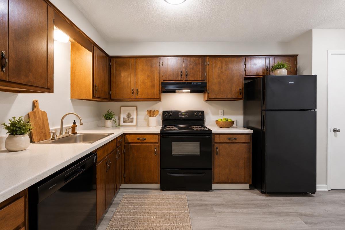 A modern kitchen featuring wooden cabinets, a black stove, and a black refrigerator. There's a light countertop with a stainless steel sink, and decorative items such as plants and kitchen utensils are visible. The flooring is a light-gray color, and the space has bright overhead lighting.