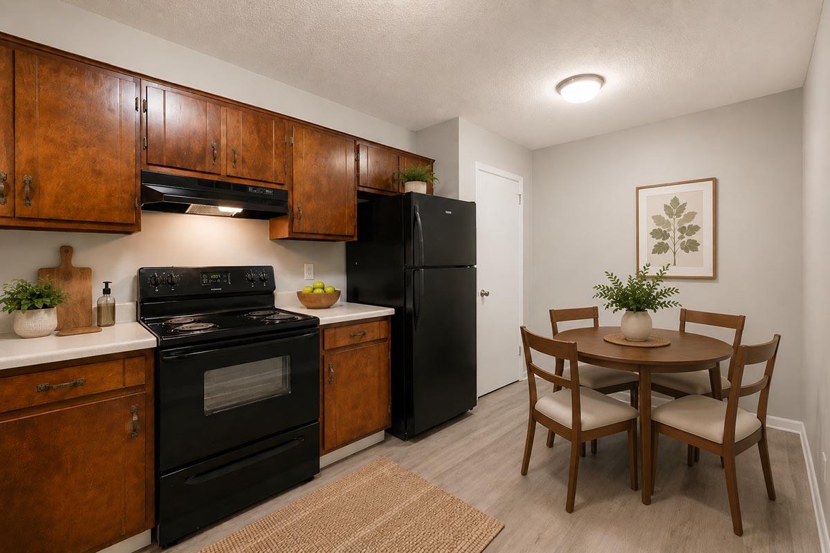 A modern kitchen featuring dark wooden cabinets, a black stove and refrigerator, and a white countertop. A small dining area with a round wooden table and four chairs is positioned nearby. A decorative plant sits on the table, and there is a framed botanical print on the wall. The floor is light-colored and the space is well-lit.