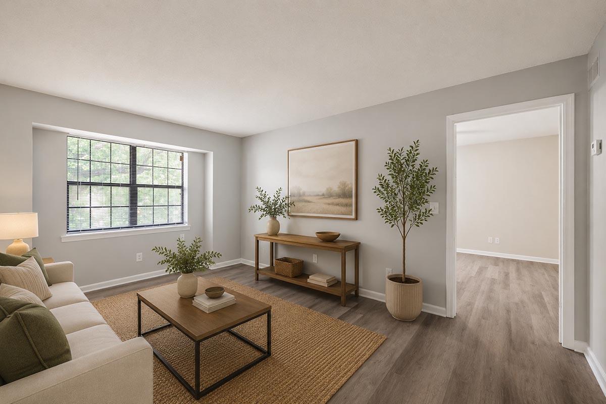 A cozy living room featuring a neutral color palette, a large window with black frames, a comfortable beige sofa adorned with green cushions, a wooden coffee table, and a simple decor piece on the wall. Potted plants add a touch of greenery, enhancing the inviting atmosphere.