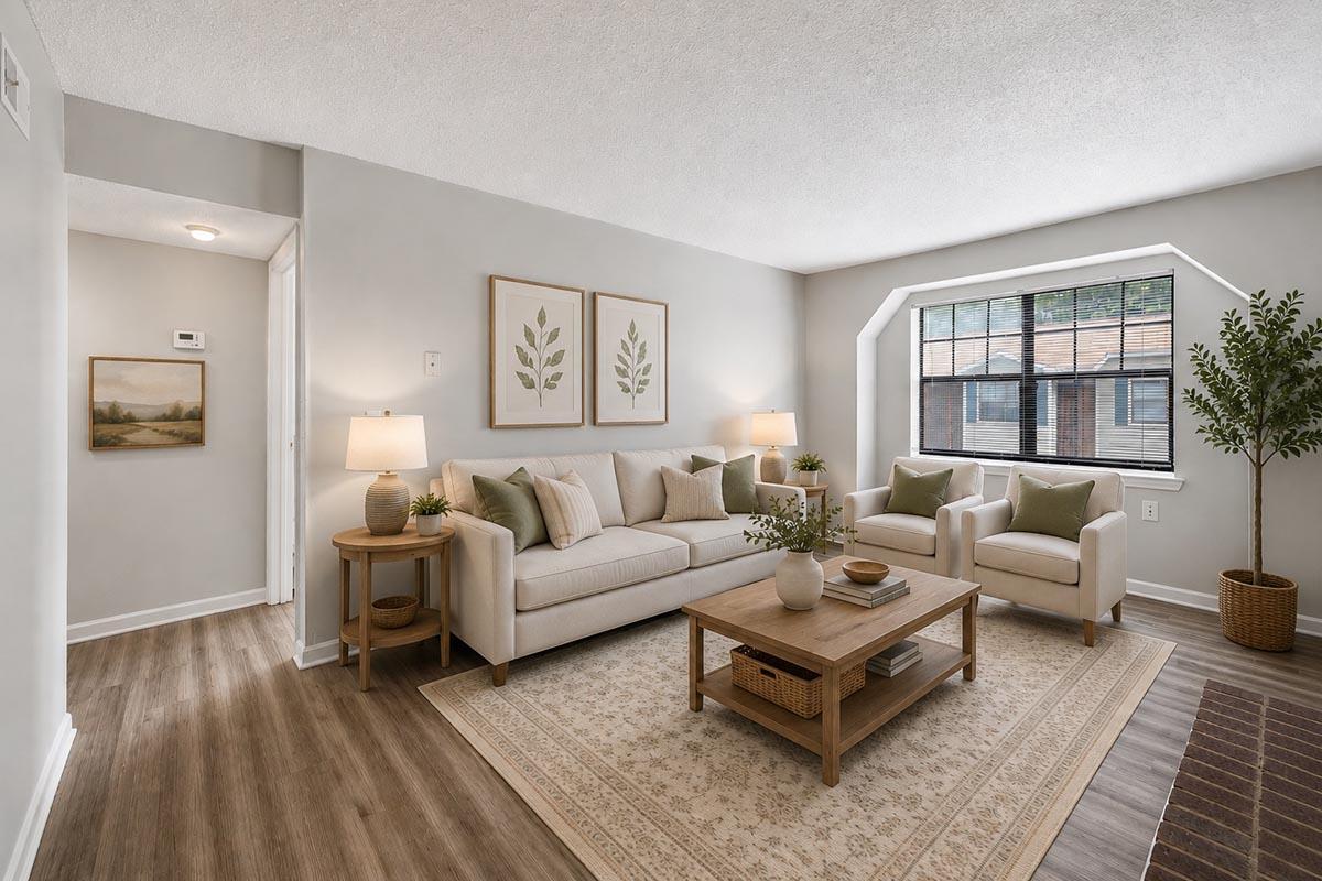 A modern living room featuring a light-colored sofa with green and beige cushions, two accent chairs, a wooden coffee table, and potted plants. The walls are painted in a soft gray, and there are framed botanical prints on the wall. A large window allows natural light to brighten the space.