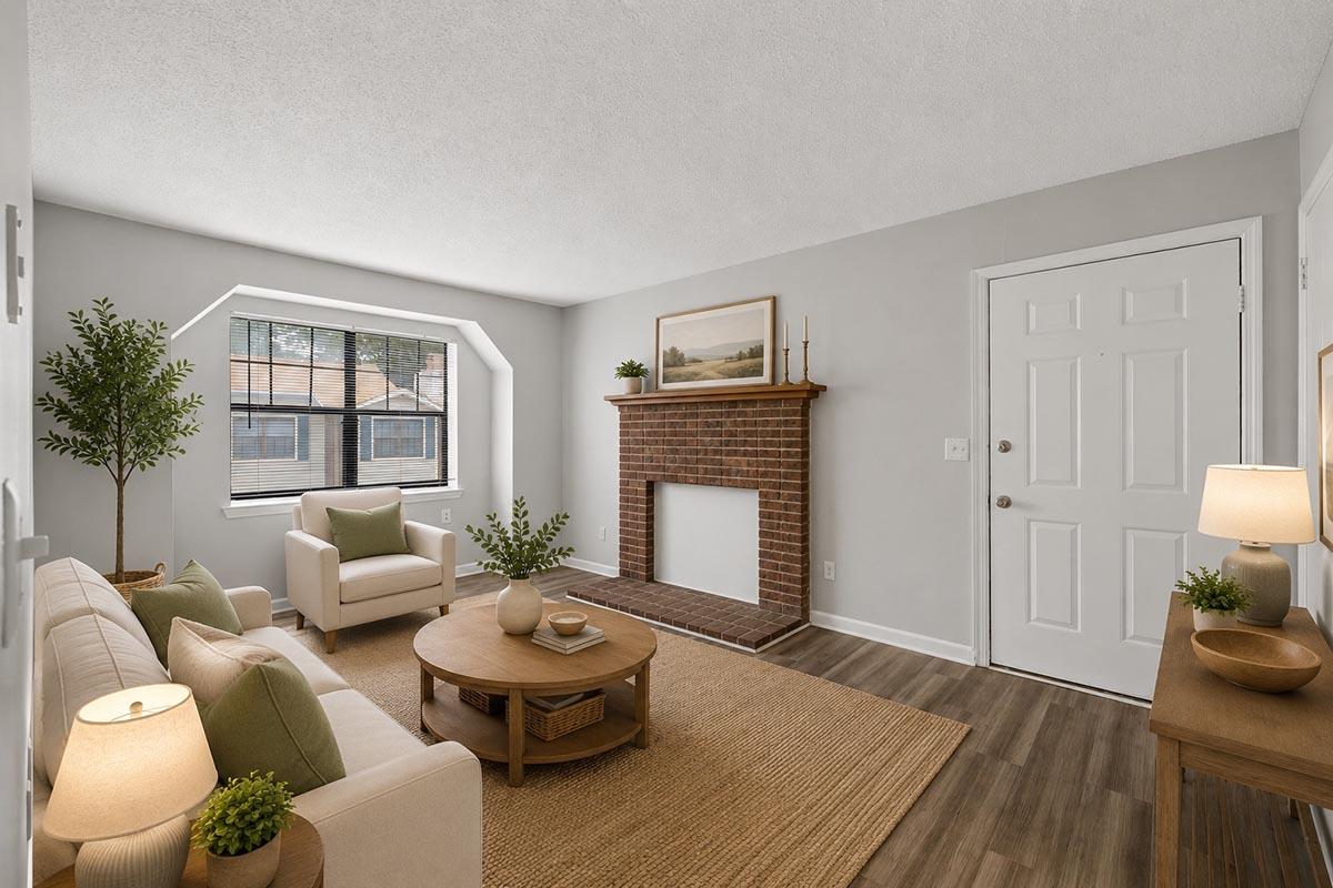 A cozy living room featuring a light-colored sofa and armchair, a round wooden coffee table, and a brick fireplace. Natural light streams in through a large window, and there are decorative plants and lamps on either side. The walls are painted in a soft gray color, creating a warm and inviting atmosphere.