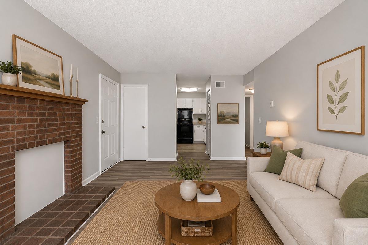 A cozy living room featuring a light-colored sofa with decorative pillows, a round coffee table, and a textured rug. There's a brick fireplace with simple decor, wall art depicting nature, and a welcoming ambiance. A doorway leads to a kitchen in the background, with soft lighting throughout the space.