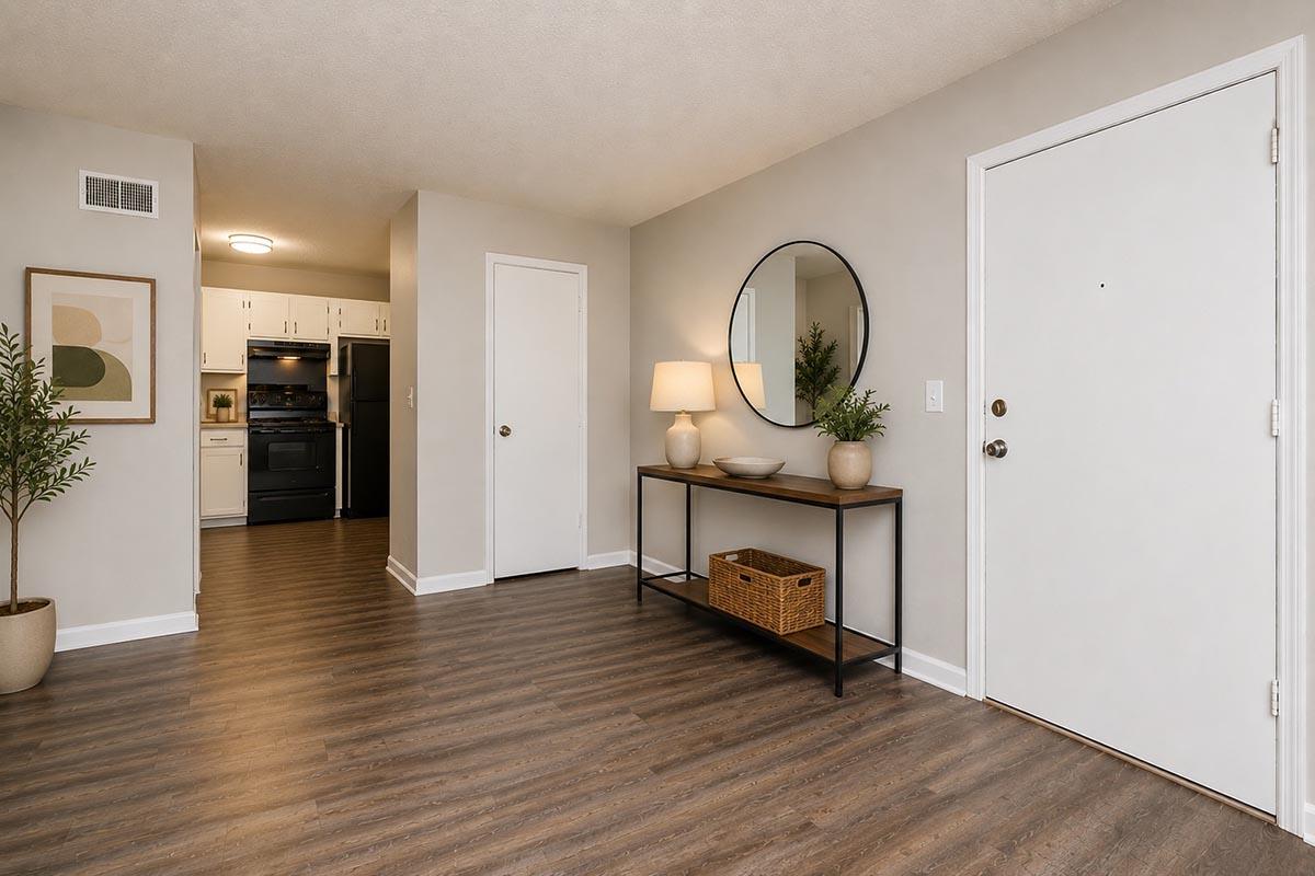A modern entryway with light gray walls, featuring a wooden table with a lamp and decorative items. A round mirror hangs above the table. To the left, a small plant in a pot adds greenery. The background shows an open kitchen area with white cabinetry and a black kitchen range, enhancing the space's contemporary feel.