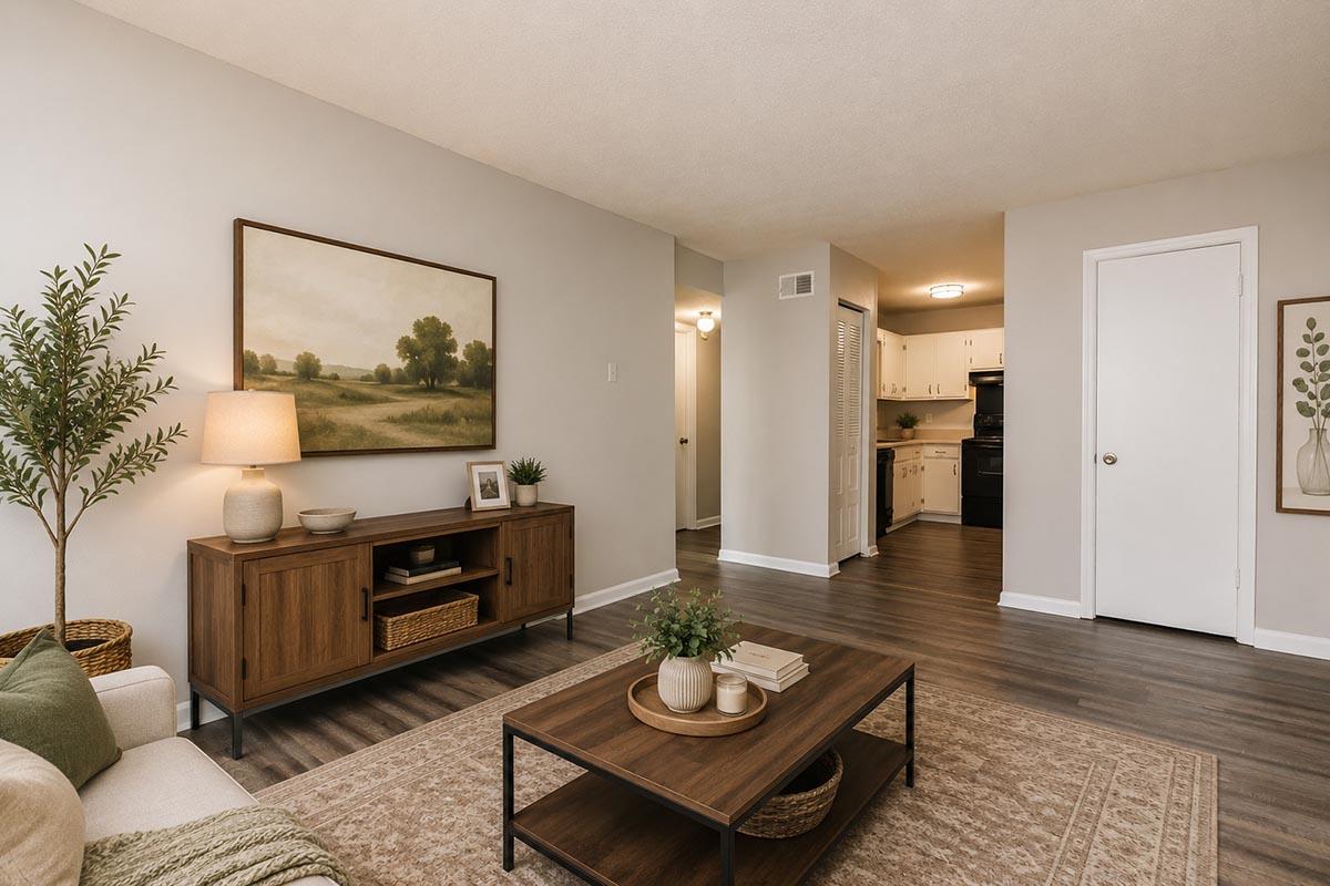 A cozy living room featuring a light-colored wall, a wooden coffee table with decor items, a beige couch, and a small tree plant. A painting of a landscape hangs on the wall, and a doorway leads to a kitchen area visible in the background. The floor has a warm wood finish, enhancing the inviting atmosphere.
