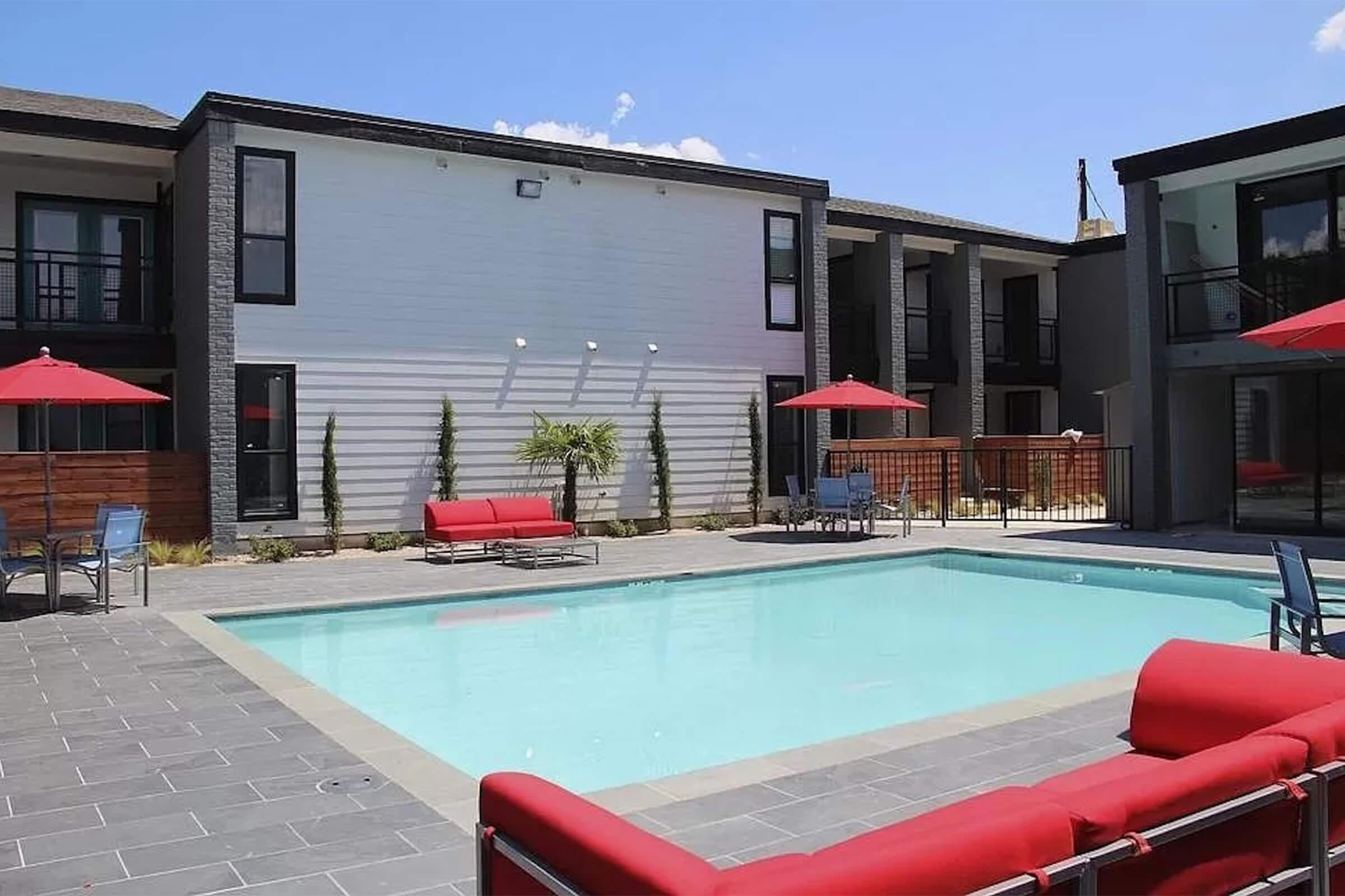A modern outdoor pool area surrounded by lounge chairs and red umbrellas. The pool is framed by contemporary buildings with a mix of brick and stucco, featuring green plants for decoration. Clear blue skies add to the inviting atmosphere of leisure and relaxation.