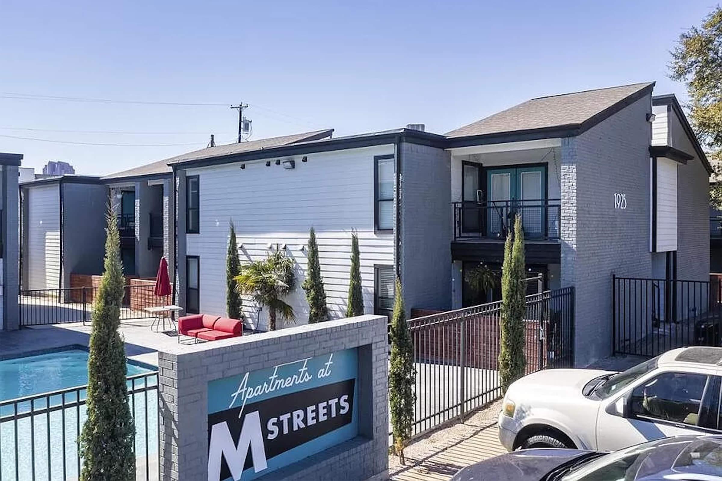 A modern apartment complex featuring a pool, landscaped greenery, and designated parking. The building has a mix of dark brick and white siding, with balconies visible on the second floor. A sign in front indicates it is "Apartments at M Streets." Sunny weather enhances the inviting atmosphere.
