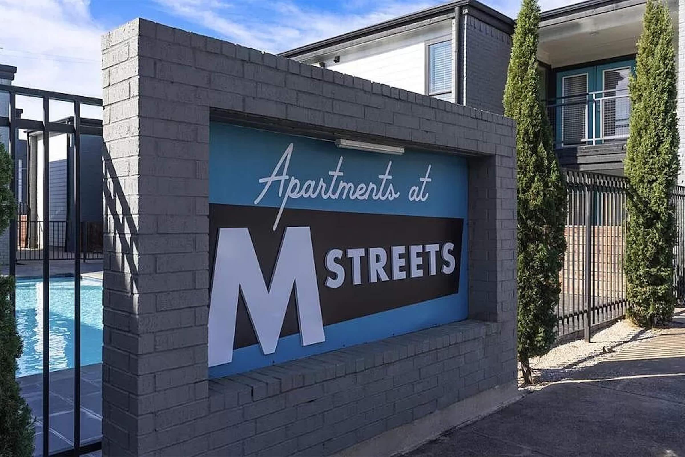 Sign for "Apartments at M Streets" with a swimming pool visible behind the fence. Surrounding the sign are tall, slender shrubs, and there are modern apartment buildings in the background under a clear blue sky. The overall scene suggests a residential community with amenities.