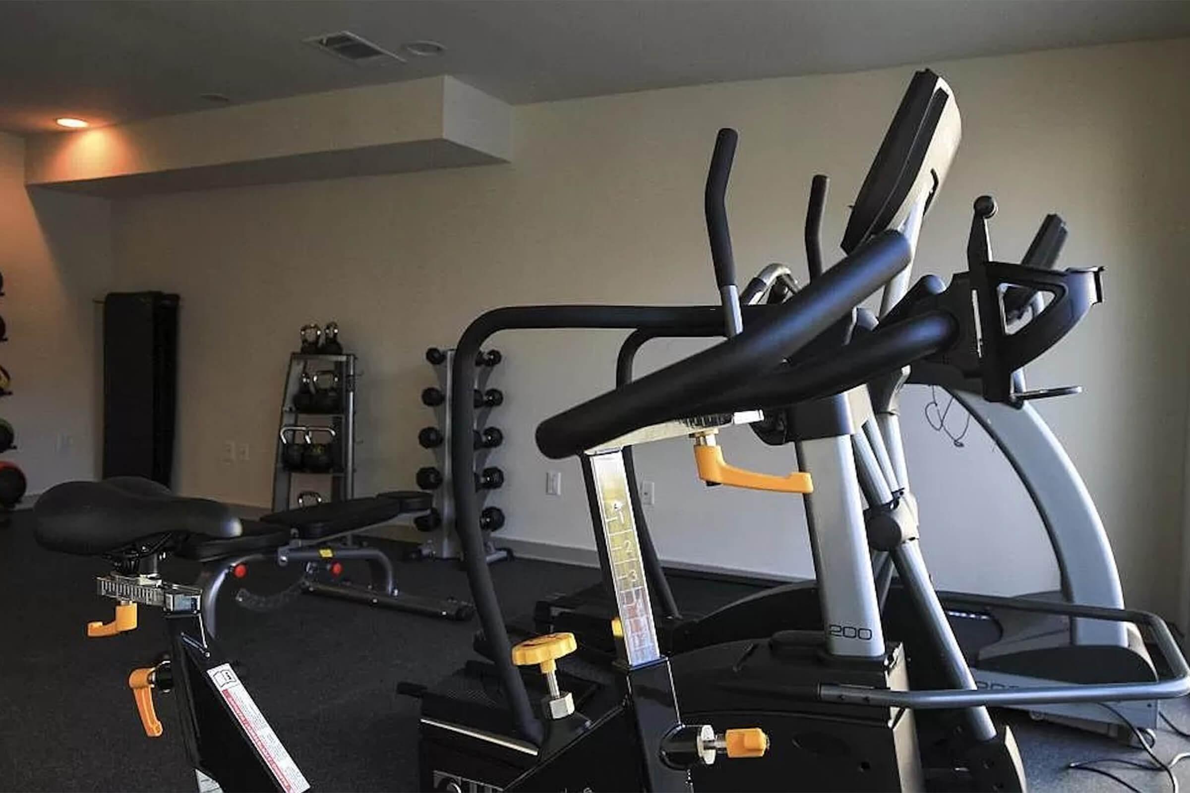 A modern home gym setup featuring an elliptical machine and a stationary bike in the foreground. In the background, there's a rack of free weights and a wall-mounted mirror, creating a spacious workout environment with neutral-colored walls and carpeted flooring.