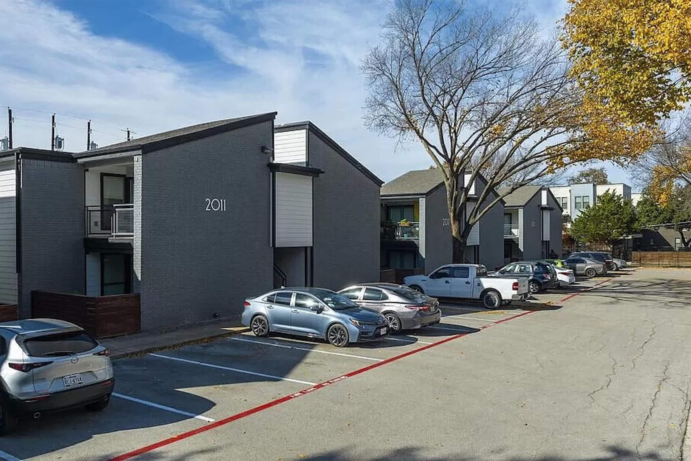 Image of an apartment complex featuring several two-story buildings. The foreground shows a parking lot with multiple parked cars, along with trees showing signs of autumn foliage. The building in focus is numbered "2011." Blue sky with scattered clouds can be seen in the background.