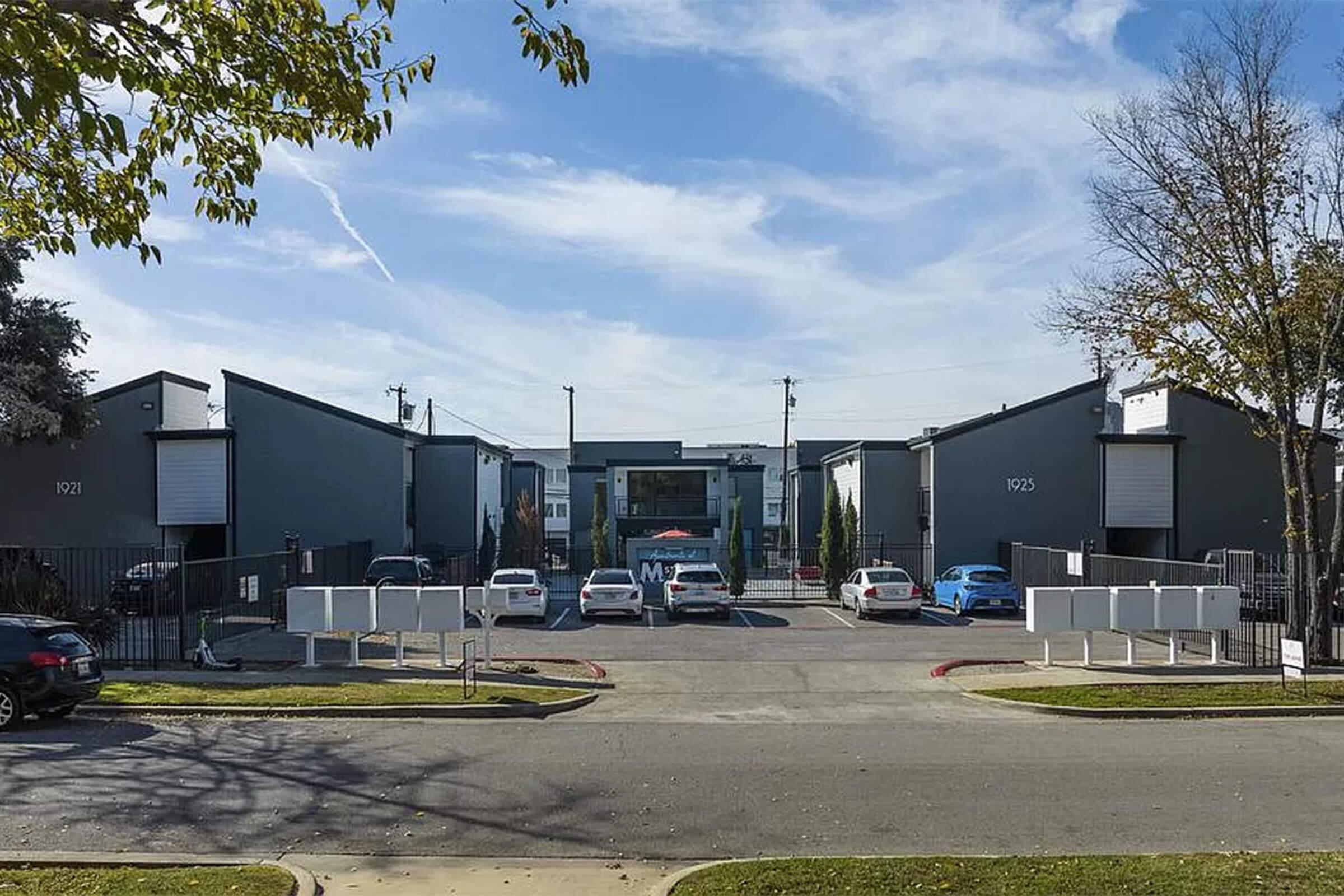 View of a residential apartment complex with a central entrance. The buildings are two stories and painted in shades of gray. Several parked cars are visible in the foreground, and there are trees lining the street. The sky is blue with scattered clouds.