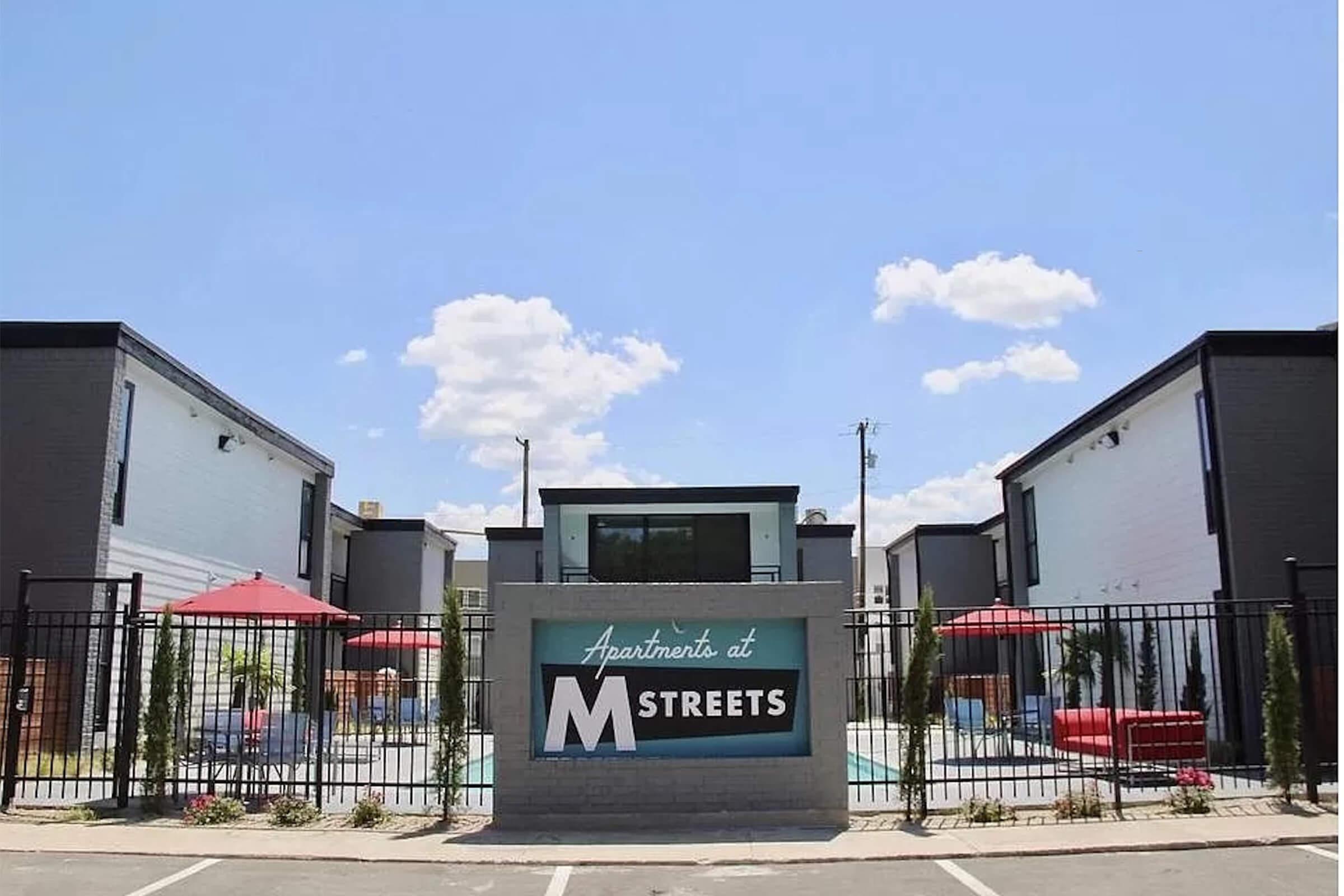 A view of the "Apartments at M Streets" complex, featuring modern buildings in a gated area. There are several trees and outdoor seating areas with red umbrellas near a pool. The sky is bright blue with a few fluffy clouds.