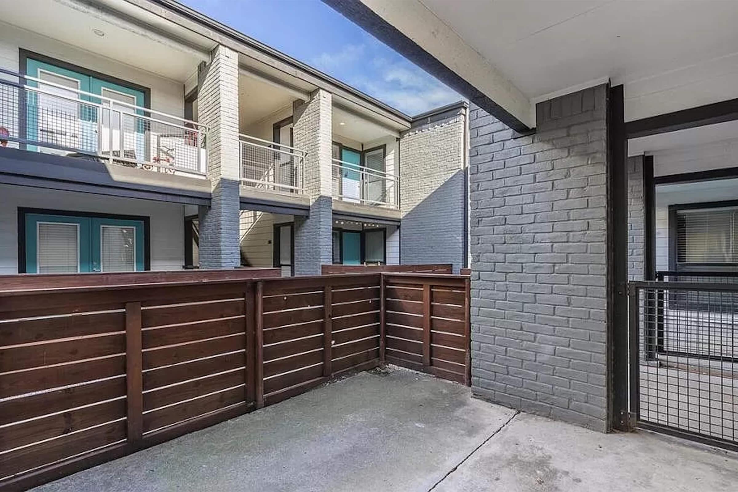 A view of a courtyard area in an apartment complex, featuring wooden fencing, a concrete floor, and surrounding brick walls. The scene includes multiple levels of apartments with balconies visible in the background under a clear blue sky.