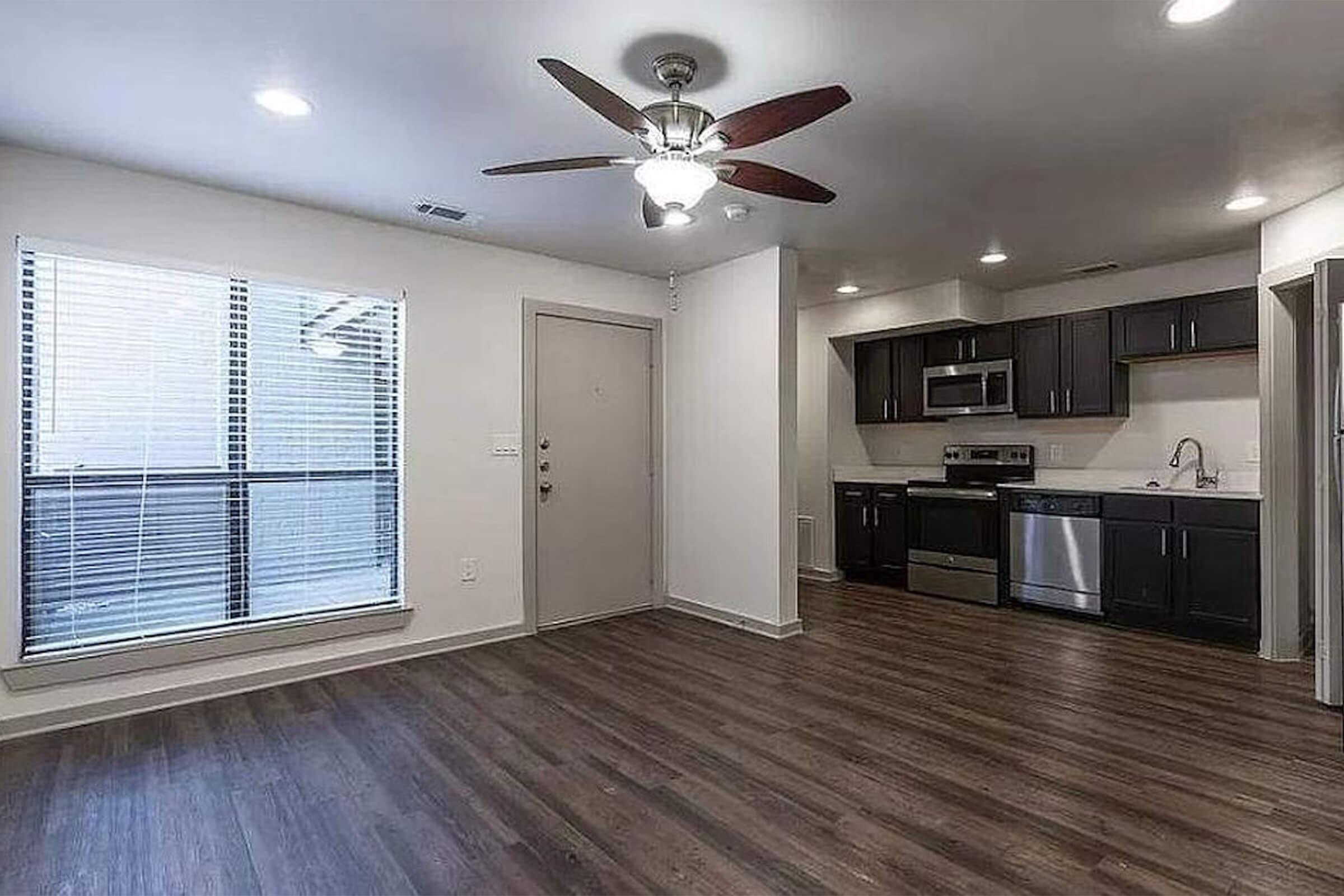 Interior view of a modern apartment featuring an open living area with hardwood flooring, a ceiling fan, and large windows. The kitchen is equipped with dark cabinets, stainless steel appliances, and a countertop. The entrance door is visible on the left, enhancing the spacious feel of the room.