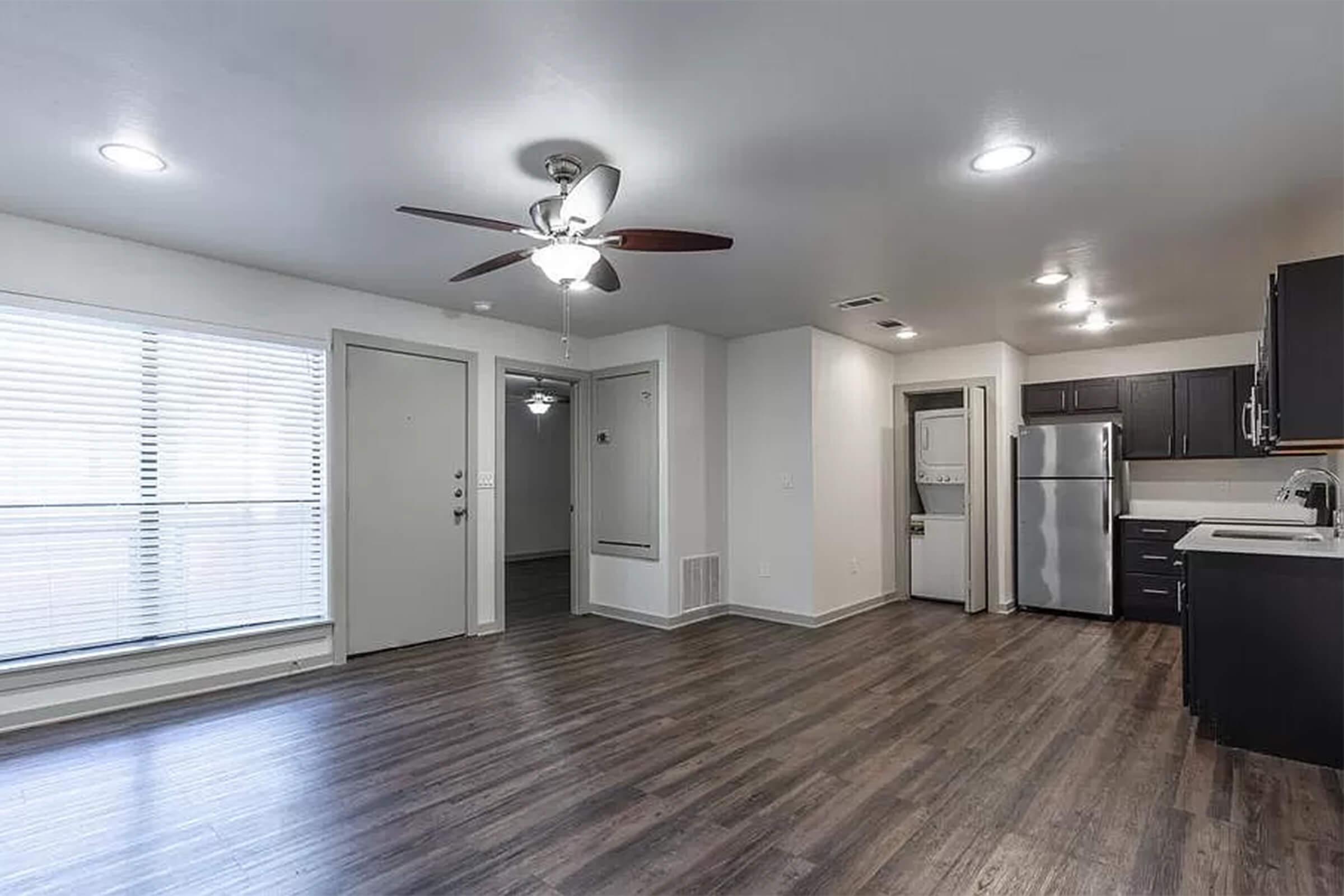 A modern, open-concept living space featuring a ceiling fan, large window with blinds, and a door leading outside. The kitchen includes stainless steel appliances and dark cabinetry. The flooring is a warm wood-like laminate, and there are light fixtures throughout the area, creating a bright and inviting atmosphere.
