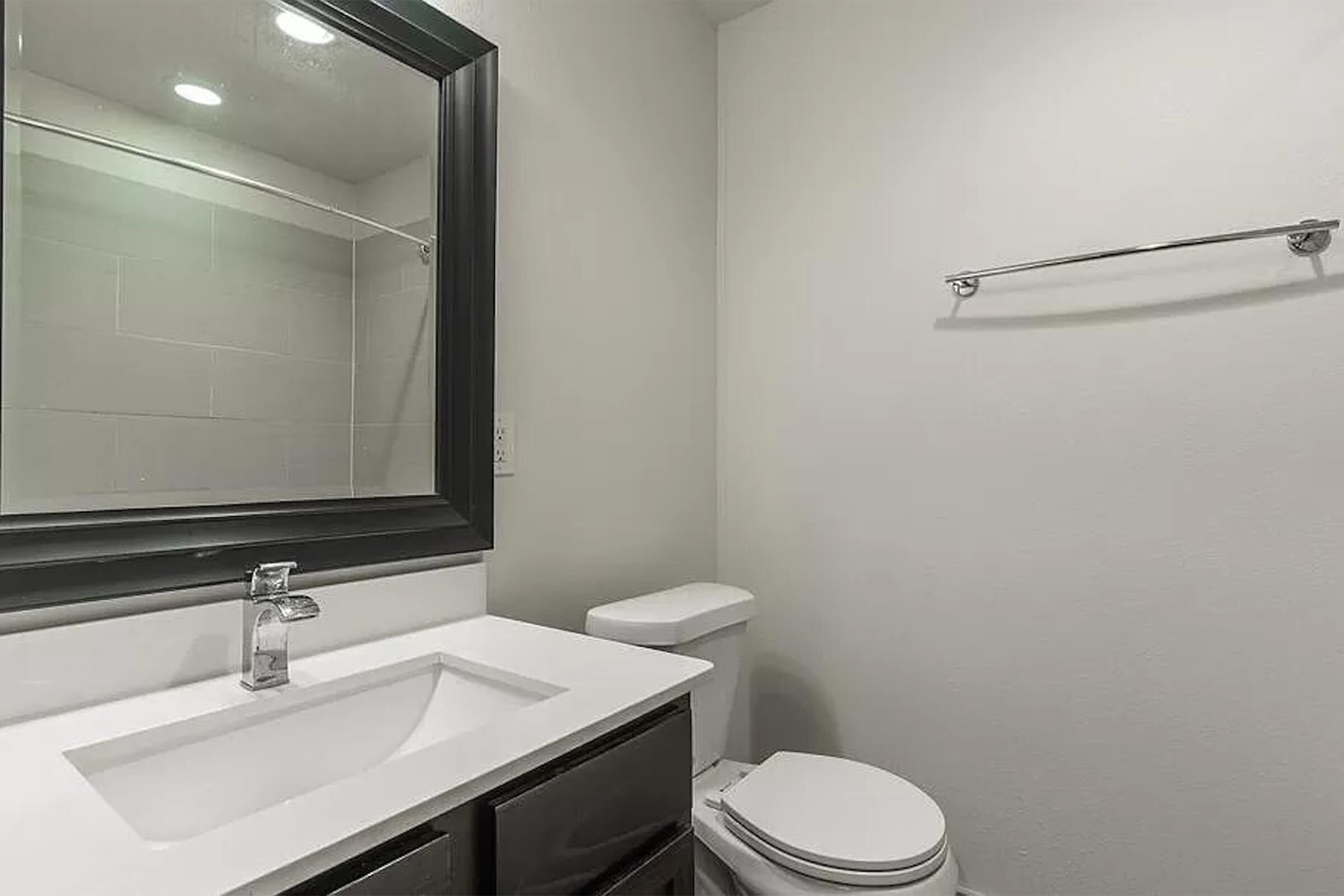 A modern bathroom featuring a sleek white countertop with a sink, a large mirror framed in black, a toilet, and a towel rack on the wall. The walls are painted in a light color, and there are recessed lights providing a bright atmosphere. The space is clean and minimalistic.