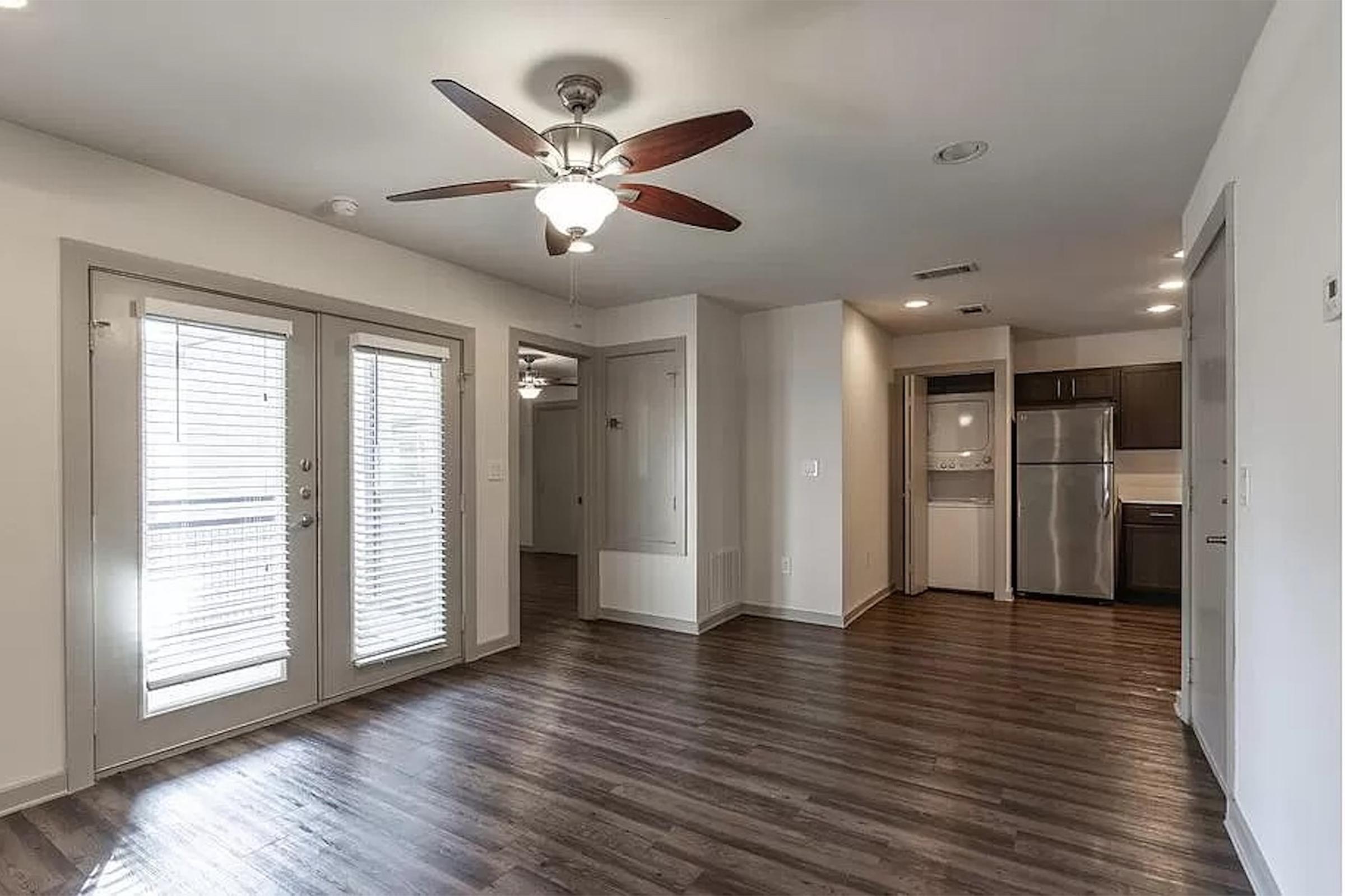 Spacious interior of a modern apartment featuring a living area with wood flooring, a ceiling fan, and sliding glass doors leading to a balcony. A kitchen area with stainless steel appliances is visible in the background, showcasing contemporary cabinetry and fixtures.