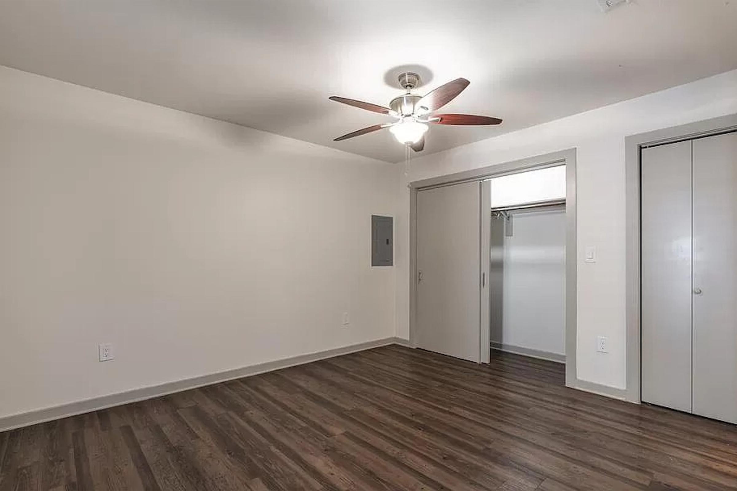 A well-lit room featuring a ceiling fan, light-colored walls, and dark wood flooring. There is a closet with sliding doors on the right side and a small panel on the left wall. The space appears clean and minimalistic, suitable for various uses like a bedroom or office.