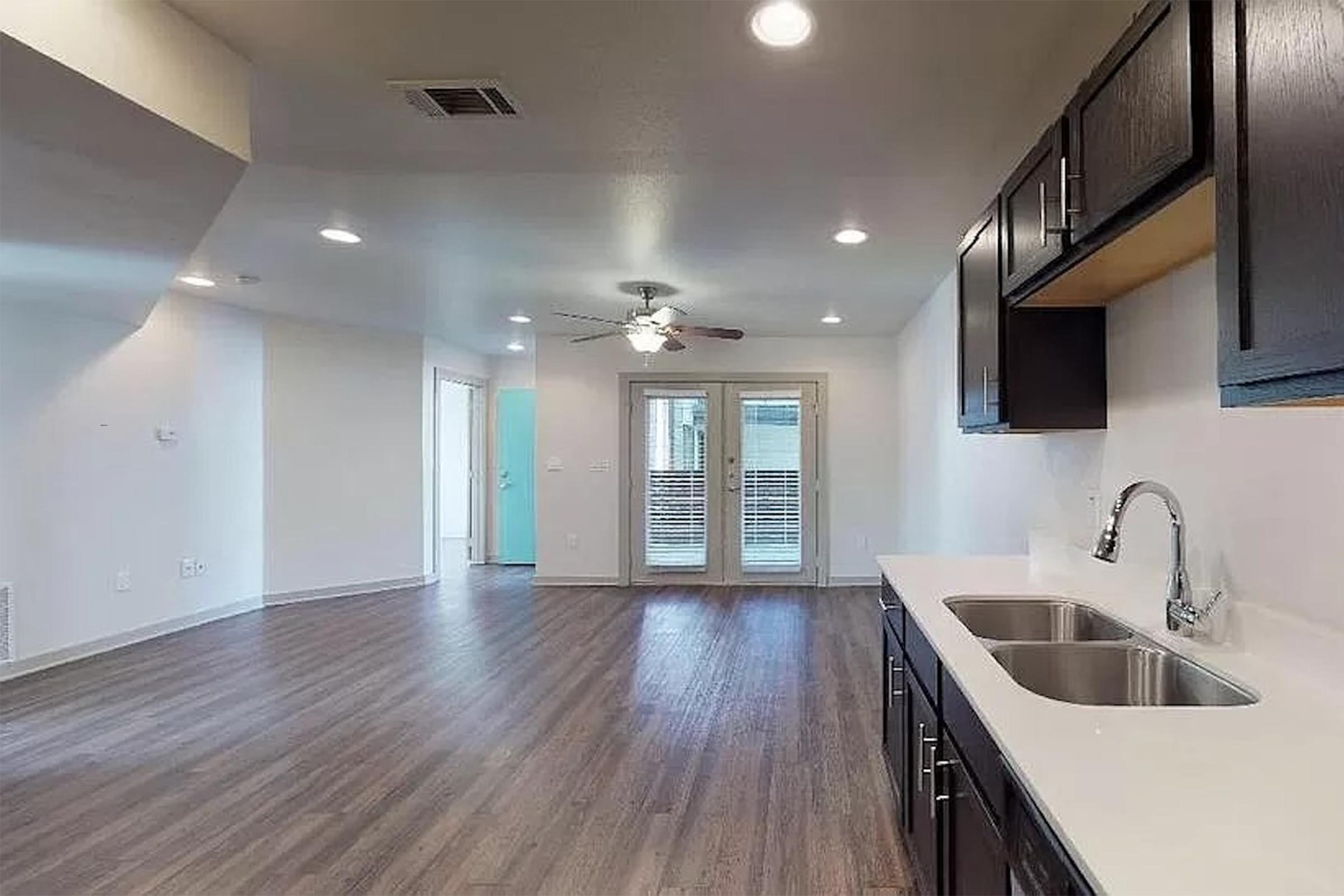 Modern kitchen and living area with light wood flooring, featuring dark cabinetry and stainless steel sink. The space has a ceiling fan and sliding glass doors leading to an outdoor area. There are bright overhead lights creating an inviting atmosphere.