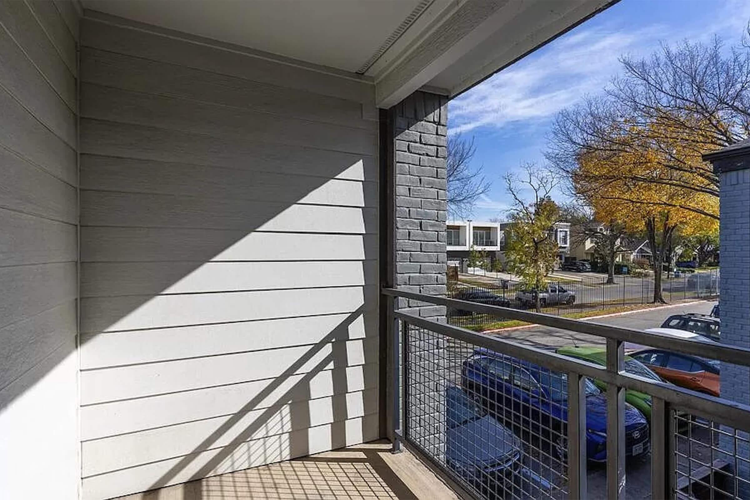 A view from a balcony featuring a gray exterior wall with a wooden railing, casting a shadow on the floor. The scene includes trees with autumn foliage in the background and a street with parked cars. The sky is clear with a few clouds, indicating a bright day.