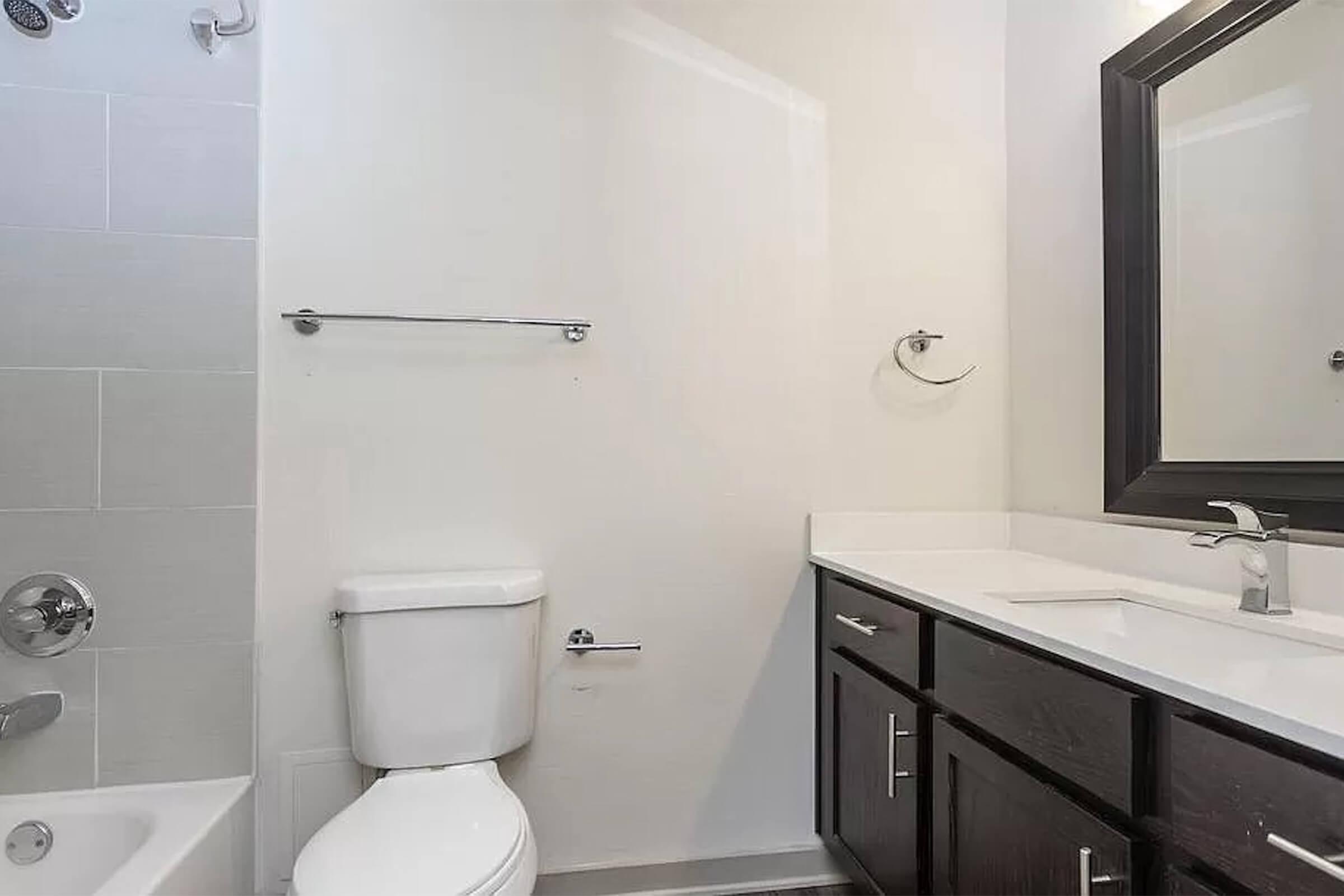 Modern bathroom featuring a white bathtub, a toilet, and a dark wood vanity with a white countertop. A large mirror is above the sink, and there are minimalistic fixtures and accessories. The walls are light gray, contributing to a clean and simple aesthetic.
