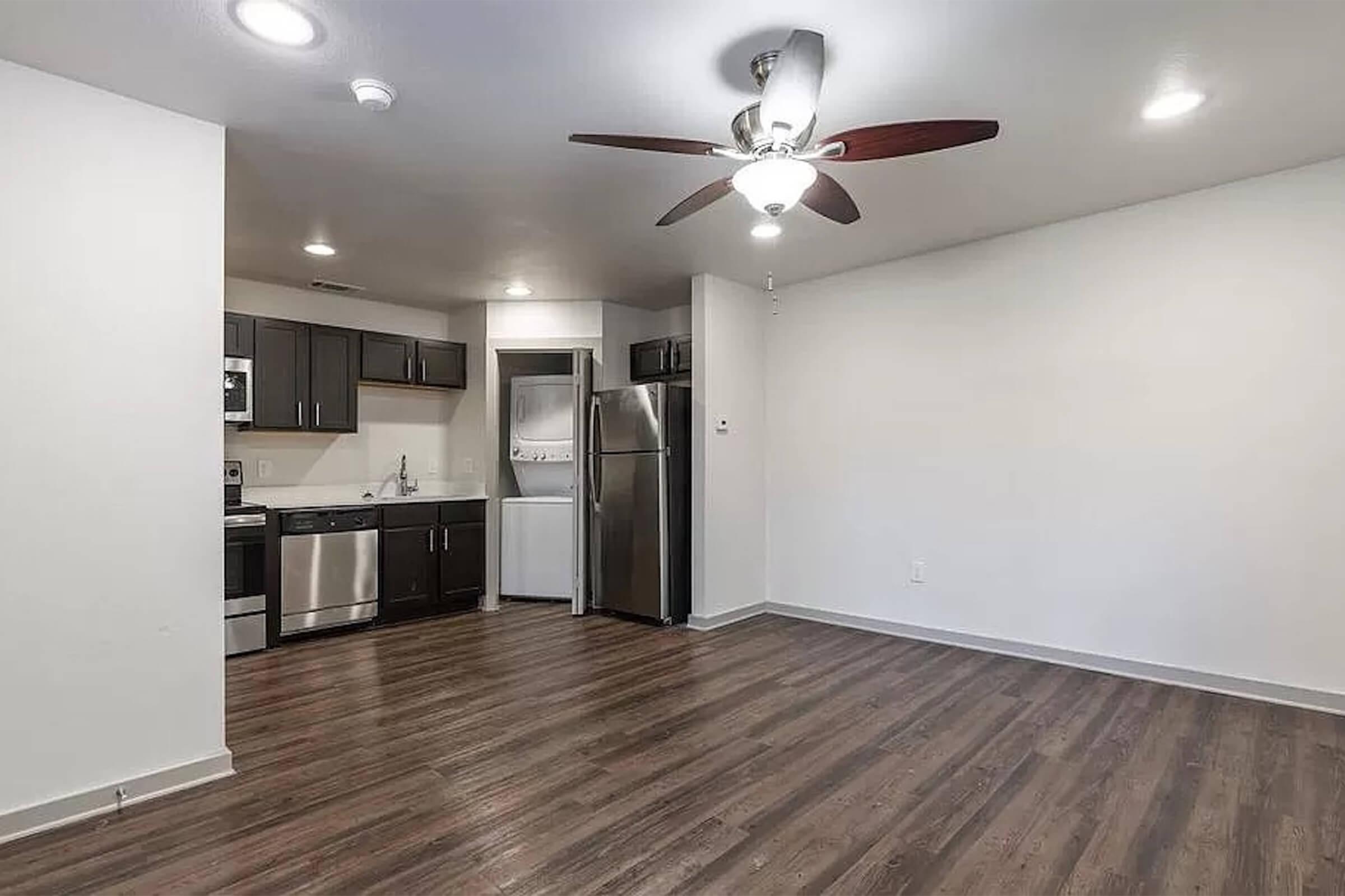 A modern, open-concept apartment interior featuring a living area with hardwood-style flooring and a ceiling fan. The kitchen is in the background, equipped with dark cabinets, stainless steel appliances, and a refrigerator. The space has neutral-colored walls and is well-lit with natural lighting.
