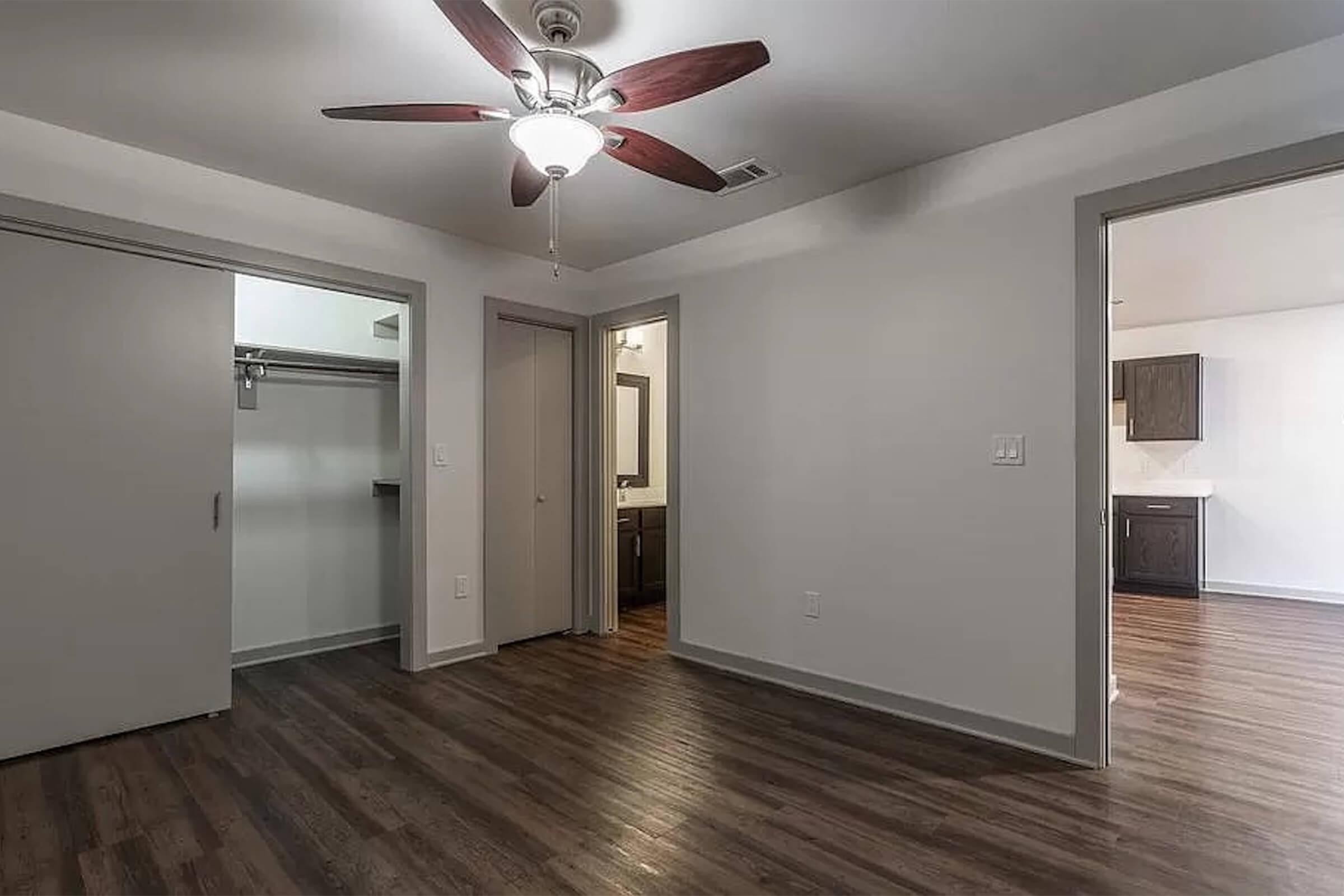 A spacious, well-lit room with a ceiling fan and wooden flooring. There are two doorways leading to other areas, one with a closet and the other to a bathroom or kitchen. The walls are painted in a light color, giving the space a modern and inviting feel.