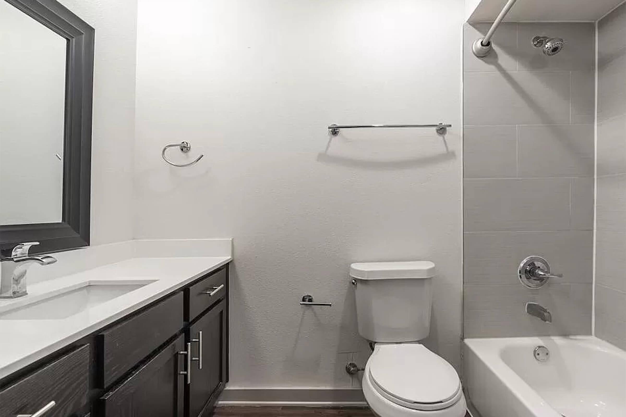 A modern bathroom featuring a rectangular mirror above a white countertop with a faucet, dark wood cabinetry below. There is a toilet beside a white bathtub with a shower fixture. The walls are light-colored tiles, and the space is well-lit with minimal decor.