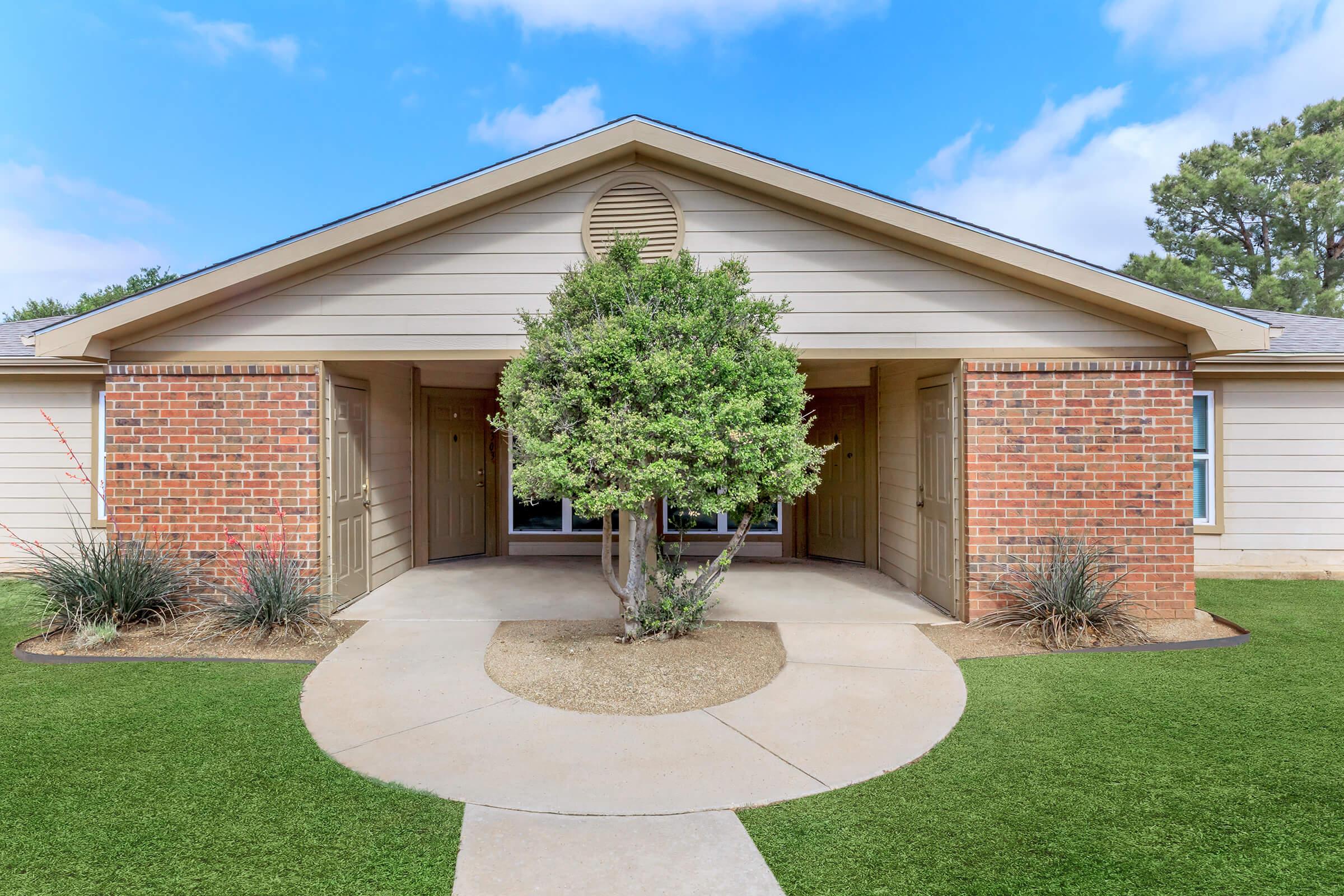 A modern single-story building with a symmetrical design, featuring a central entrance flanked by brick columns and small landscaped areas. A round concrete pathway leads to the entrance, which is partially shaded by a small tree. The building has a light-colored exterior and is set against a bright blue sky.