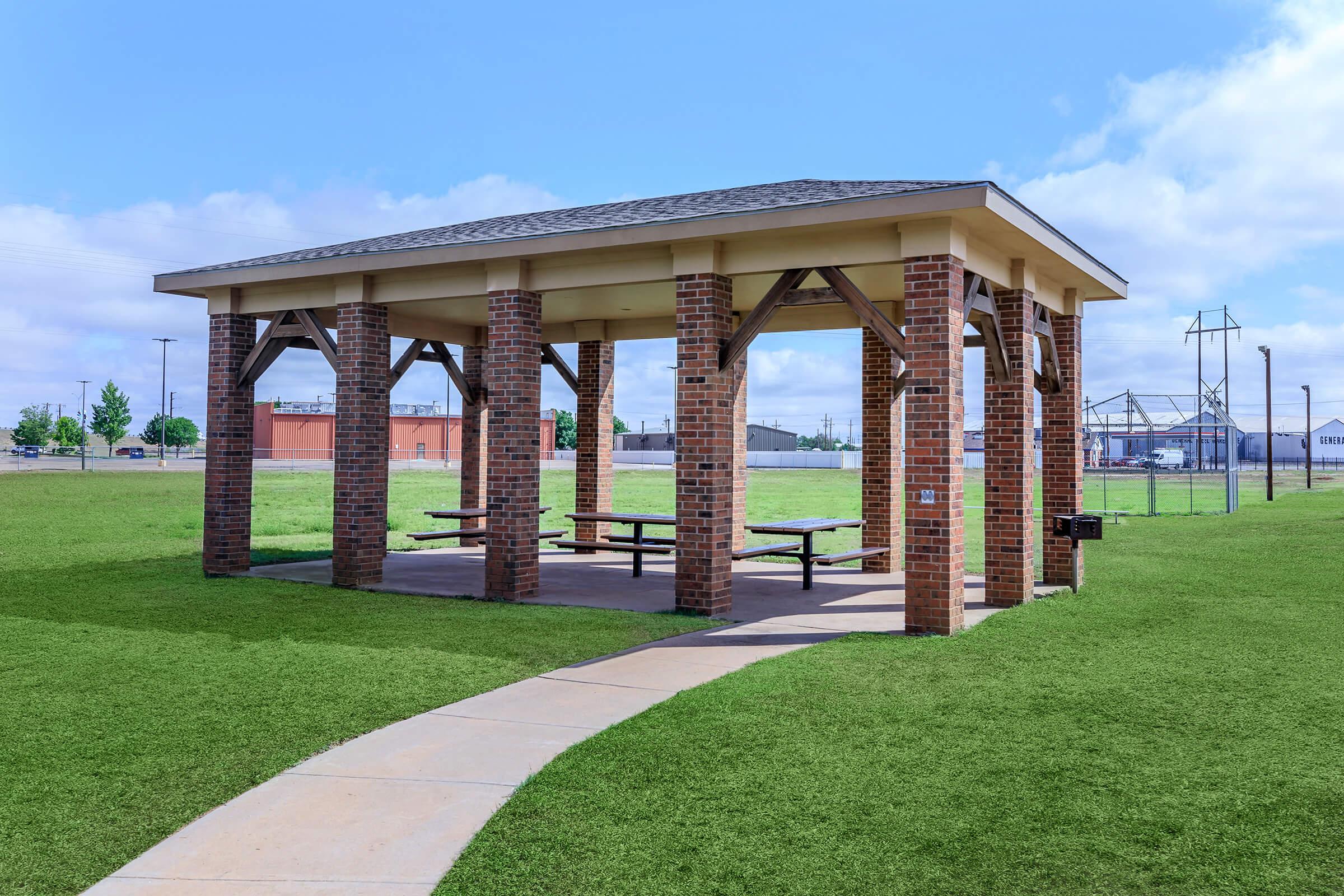 A brick pavilion with a roof supported by wooden beams, surrounded by green grass. There are picnic tables underneath, and a paved path leading to the structure. The background features a blue sky with clouds and a hint of industrial buildings in the distance.