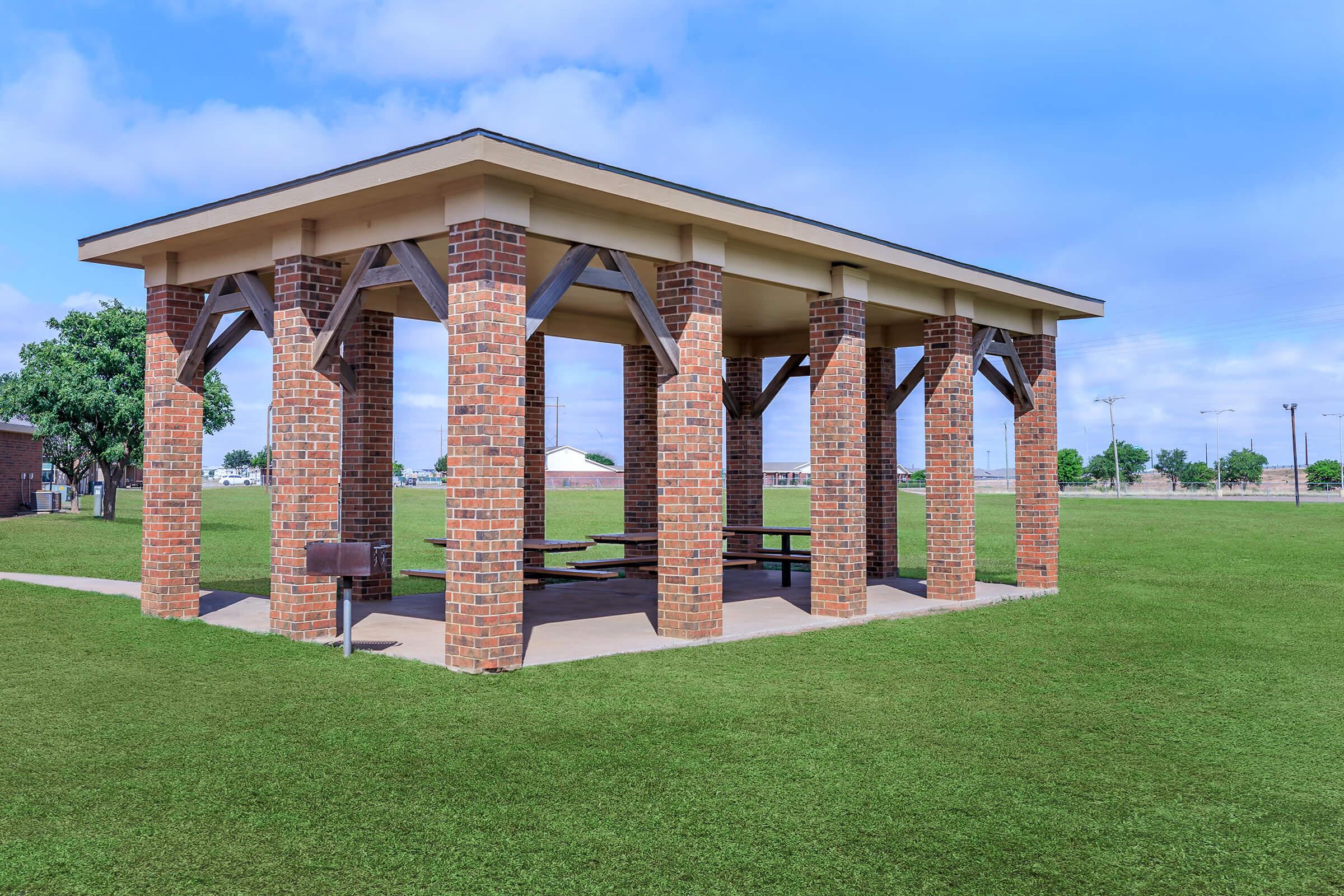 A rustic brick pavilion featuring a wooden frame, set in a spacious green park. The structure has open sides and a concrete floor, surrounded by well-maintained grass. In the background, there are a few trees and distant buildings under a bright blue sky with some clouds.