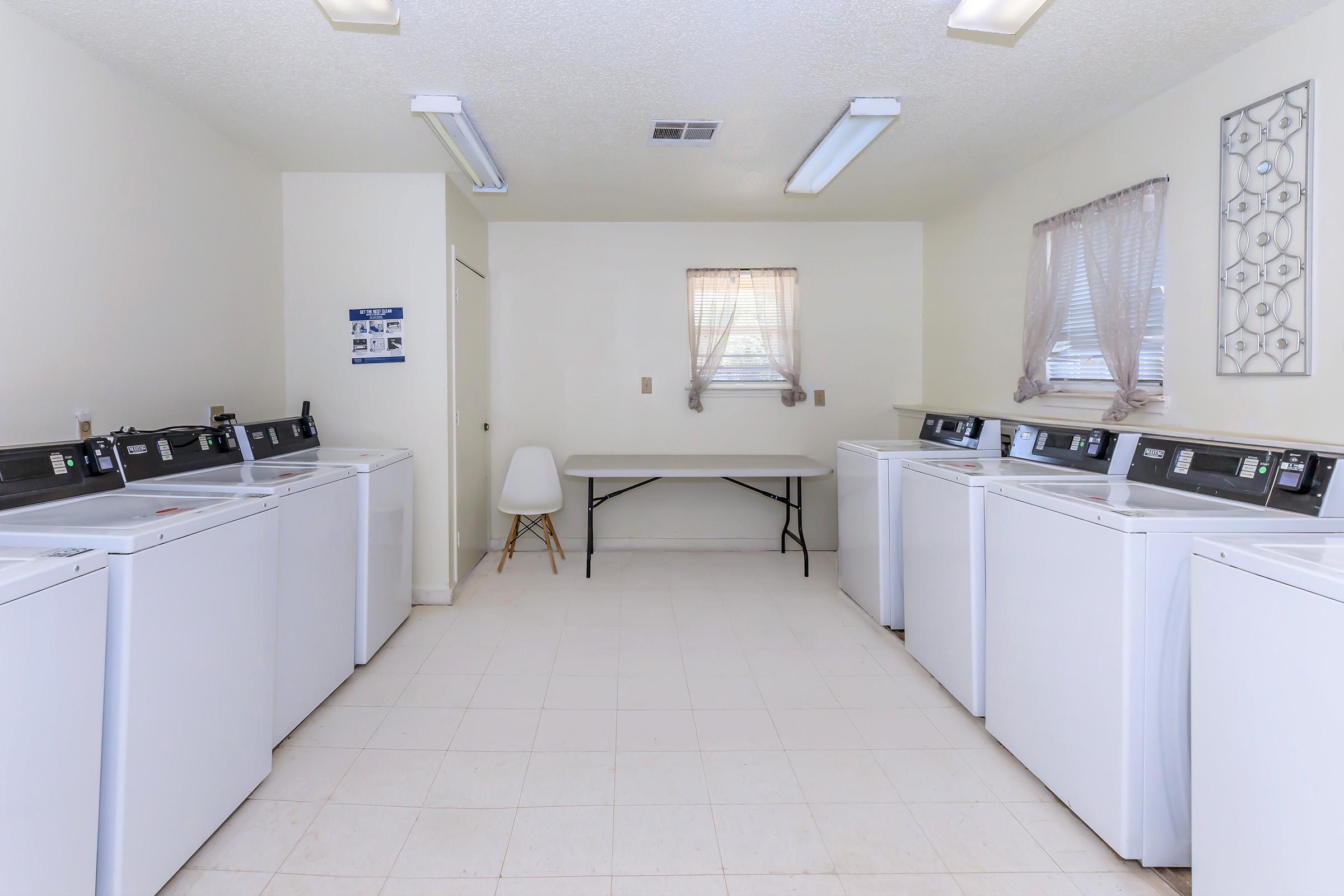 A clean, bright laundry room featuring several white washing machines and a table in the center. The walls are painted white, and there are two windows with sheer curtains allowing natural light to fill the space.