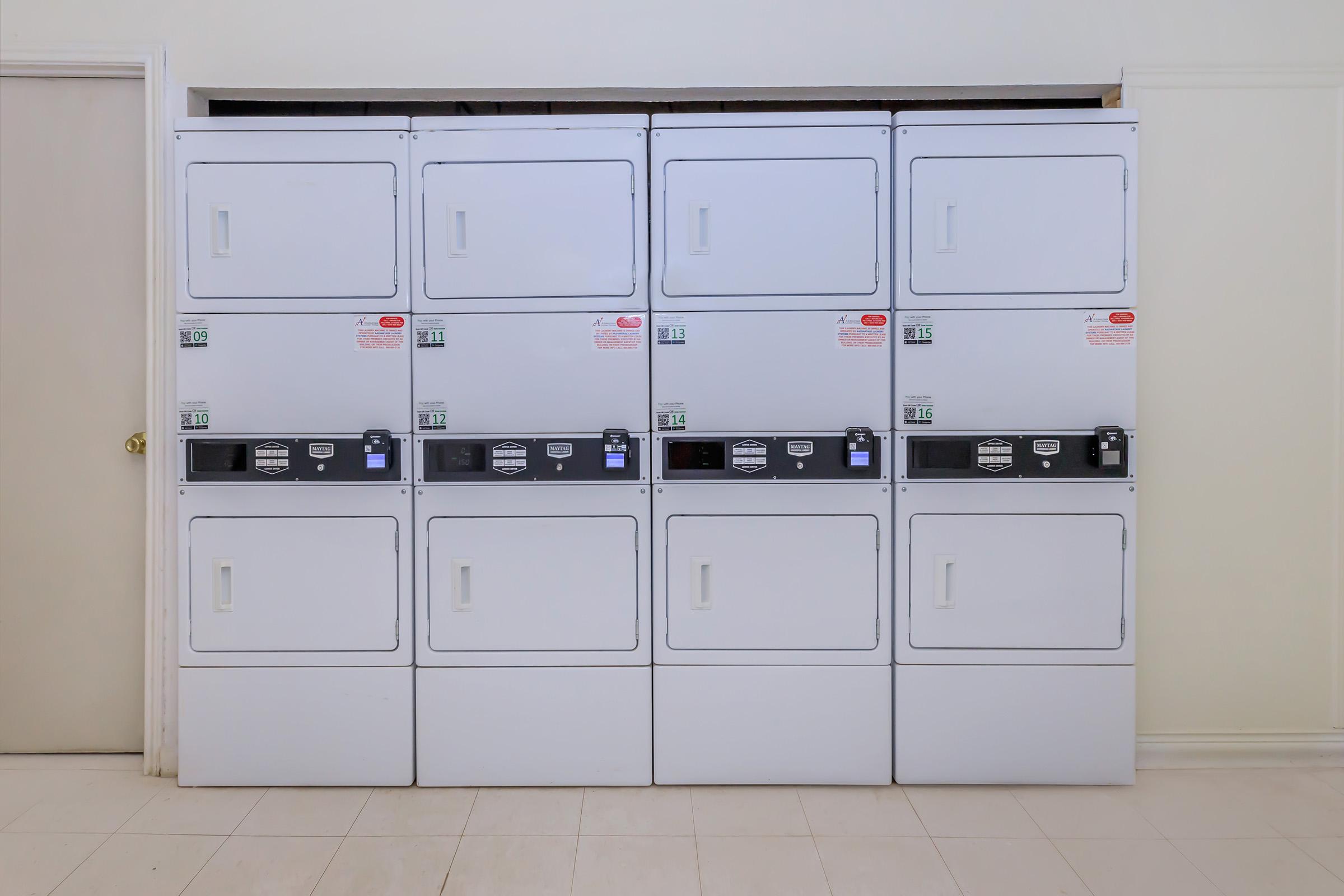 A row of white stacked laundry machines, including dryers and washers, arranged neatly against a light-colored wall. The machines feature digital displays and control panels, and there is a door adjacent to the laundry setup, contributing to a clean and functional laundry room atmosphere.