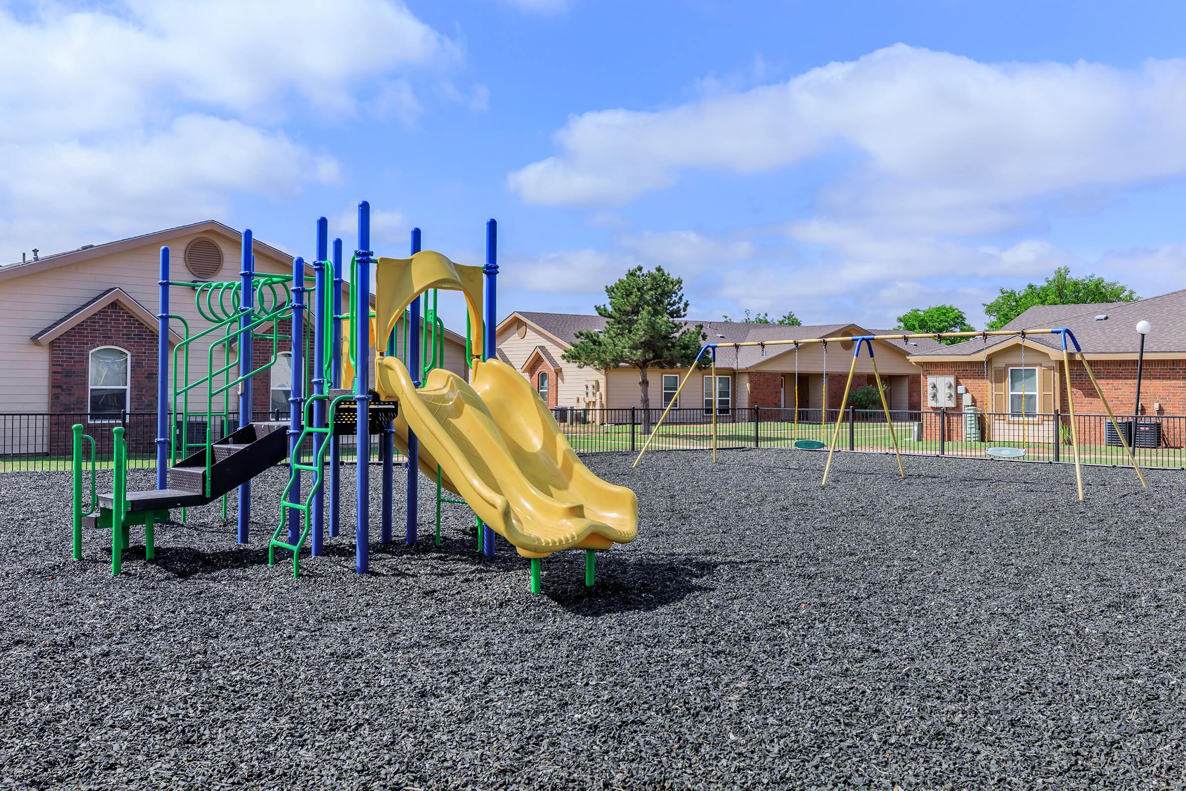A children's playground featuring a yellow slide and climbing structure, surrounded by safety surfacing. In the background, there are swings and residential buildings. The sky is partly cloudy, creating a bright atmosphere.