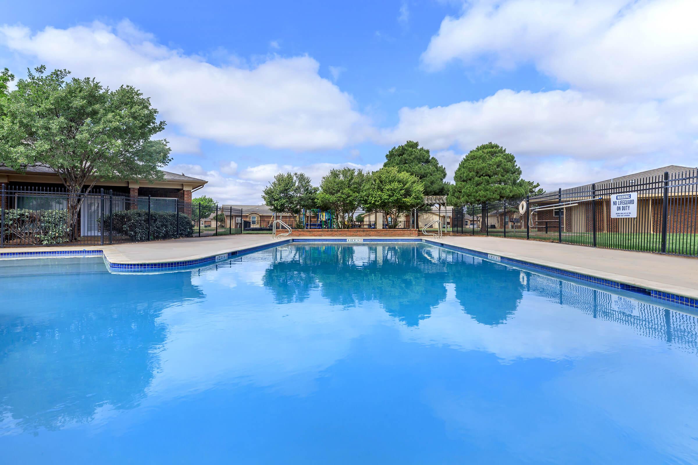A clear blue swimming pool surrounded by a landscaped area with trees and shrubs. In the background, there are buildings with a fence enclosing the pool area. The sky is partly cloudy, creating a peaceful and inviting atmosphere.