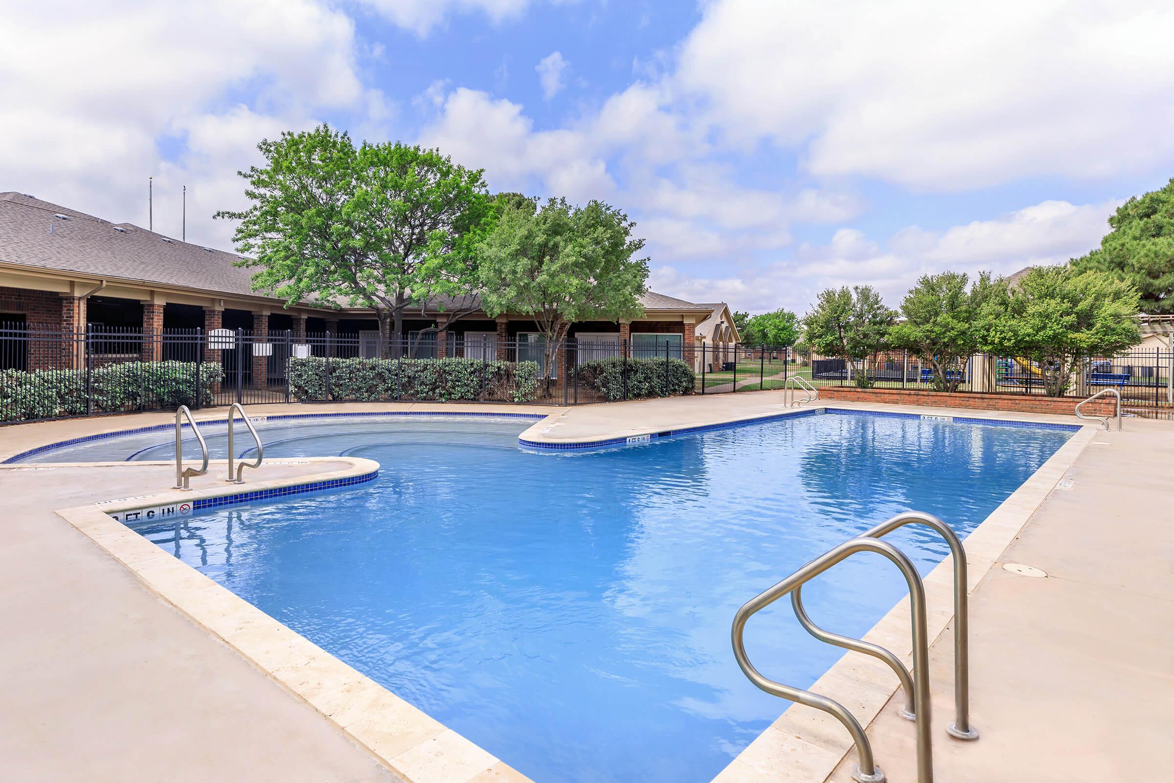 A large swimming pool with a curved edge and steps leading into the water. Surrounding the pool are landscaped areas with various trees and shrubs. The pool area is well-maintained, featuring a light-colored deck and a fence in the background, under a partly cloudy sky.