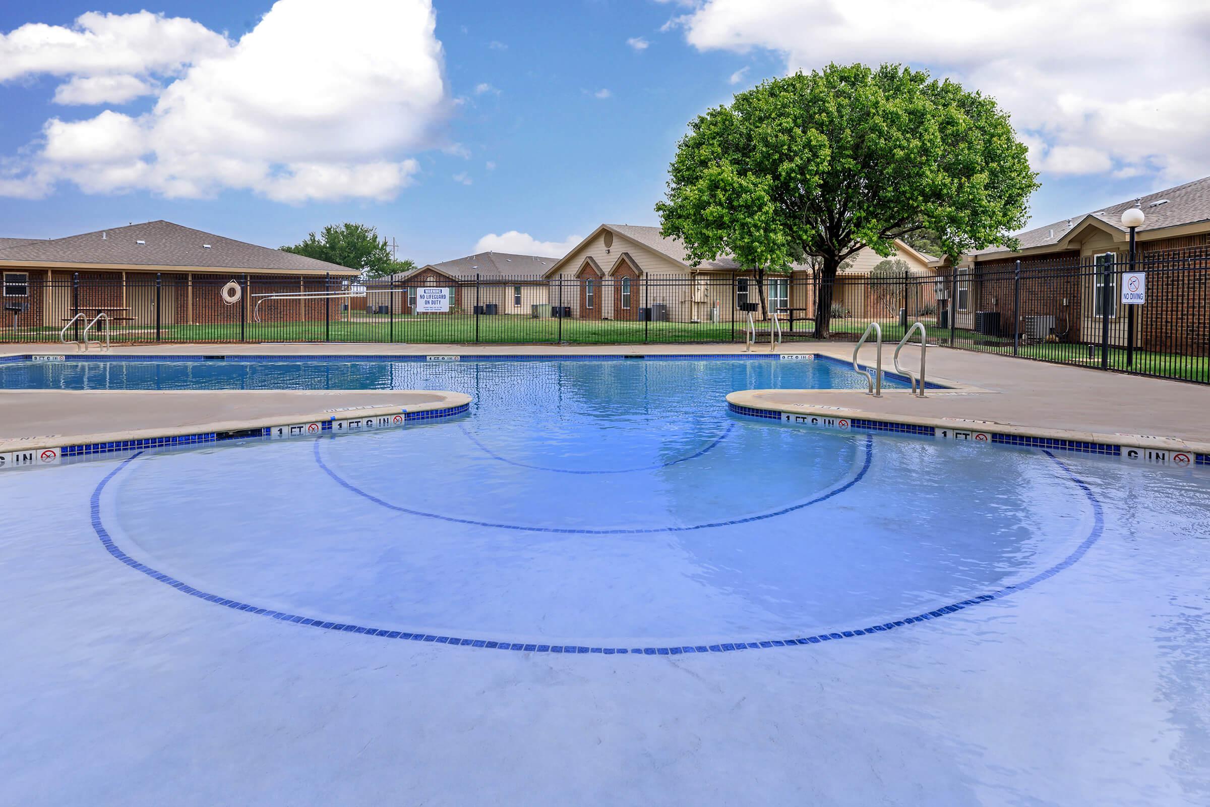 A clear swimming pool with curved edges, surrounded by a deck and grassy area. In the background, there are brick buildings with sloped roofs and a large tree. The sky is bright blue with fluffy white clouds. The scene conveys a peaceful outdoor space perfect for relaxation and recreation.