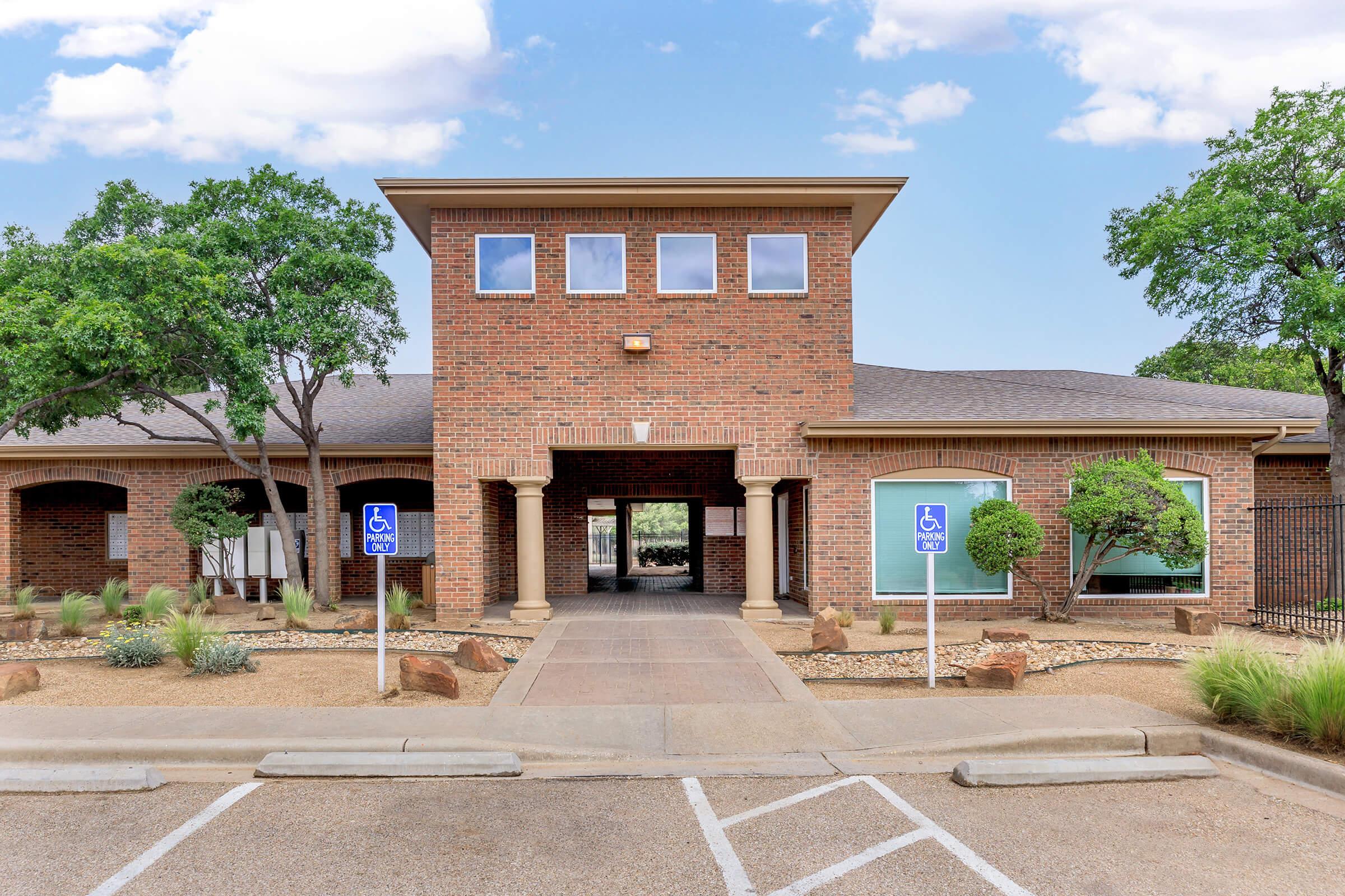 Brick building with a flat roof featuring large front windows and two accessible parking spaces in front. The landscape includes small shrubs and trees, and a pathway leads to the entrance. The sky is clear with a few clouds.