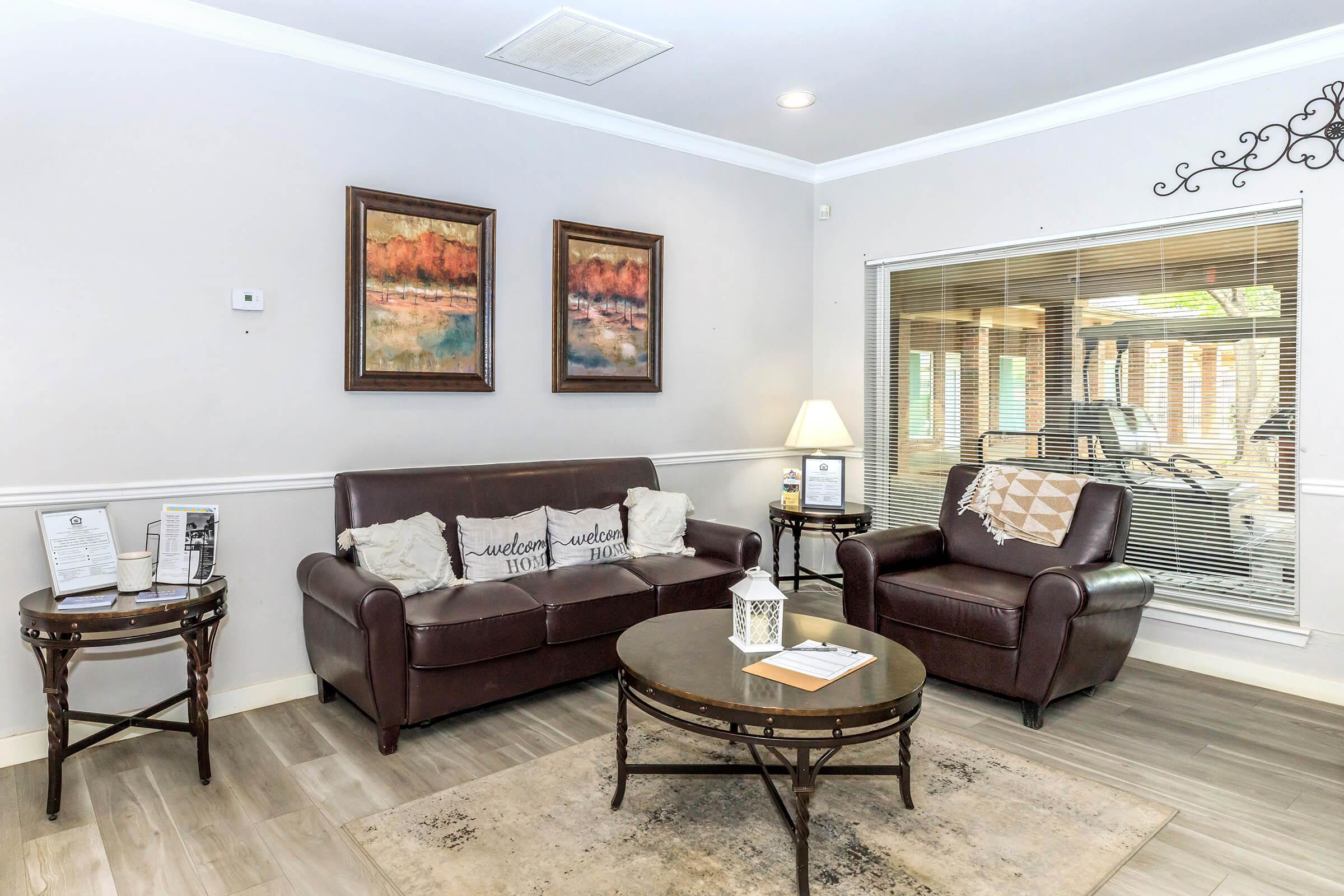 A cozy living room with two brown leather couches, a round table in the center, and decorative pillows. The walls are light-colored, adorned with two framed paintings. A lamp provides soft lighting, and a large window with blinds shows outdoor greenery. Brochures are on a side table.