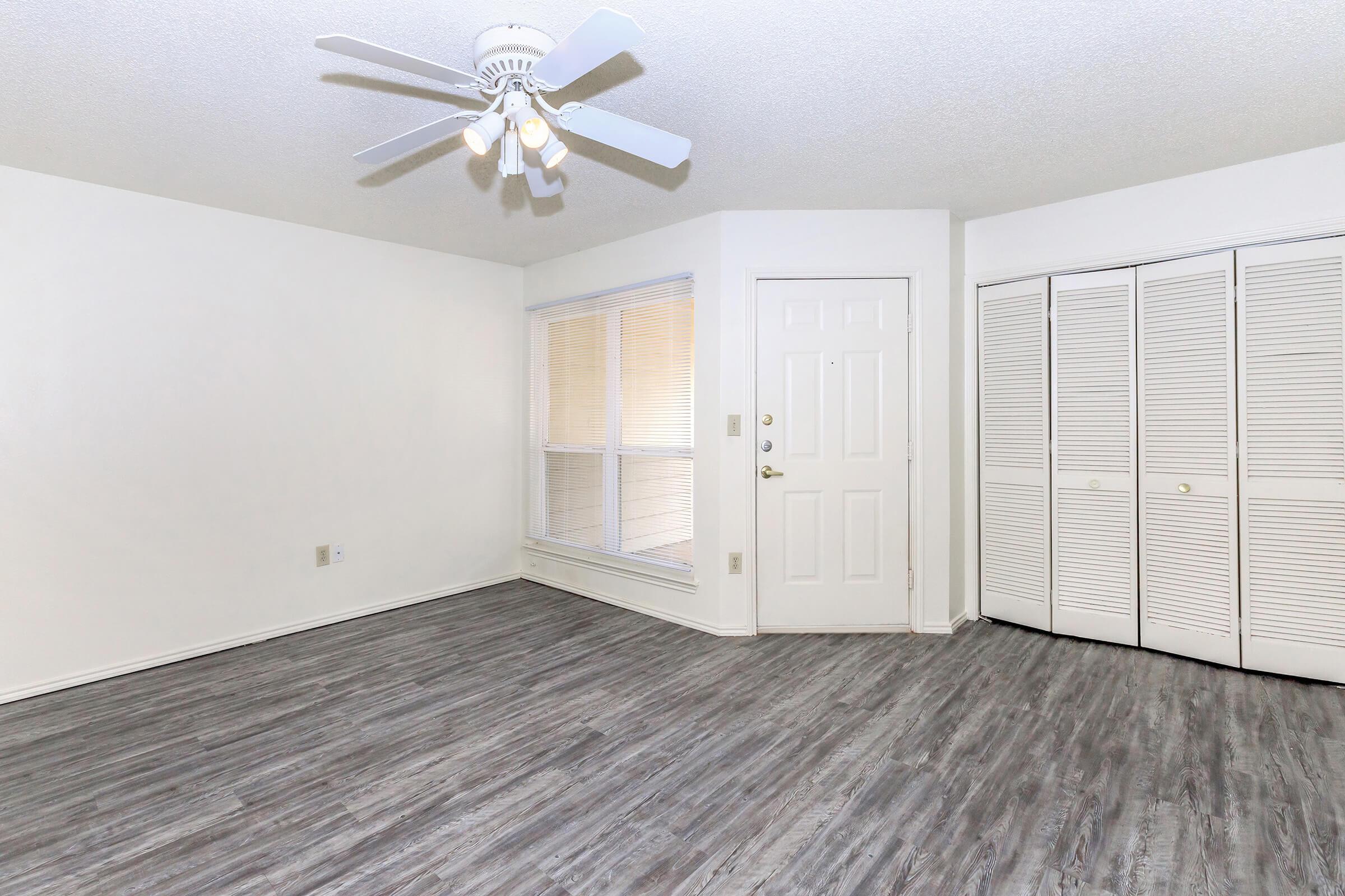 A spacious, empty room featuring light-colored walls and a ceiling fan. The floor is covered in modern, gray laminate. There's a door leading outside, large windows with blinds, and a closet with white bifold doors, creating a bright and open atmosphere.