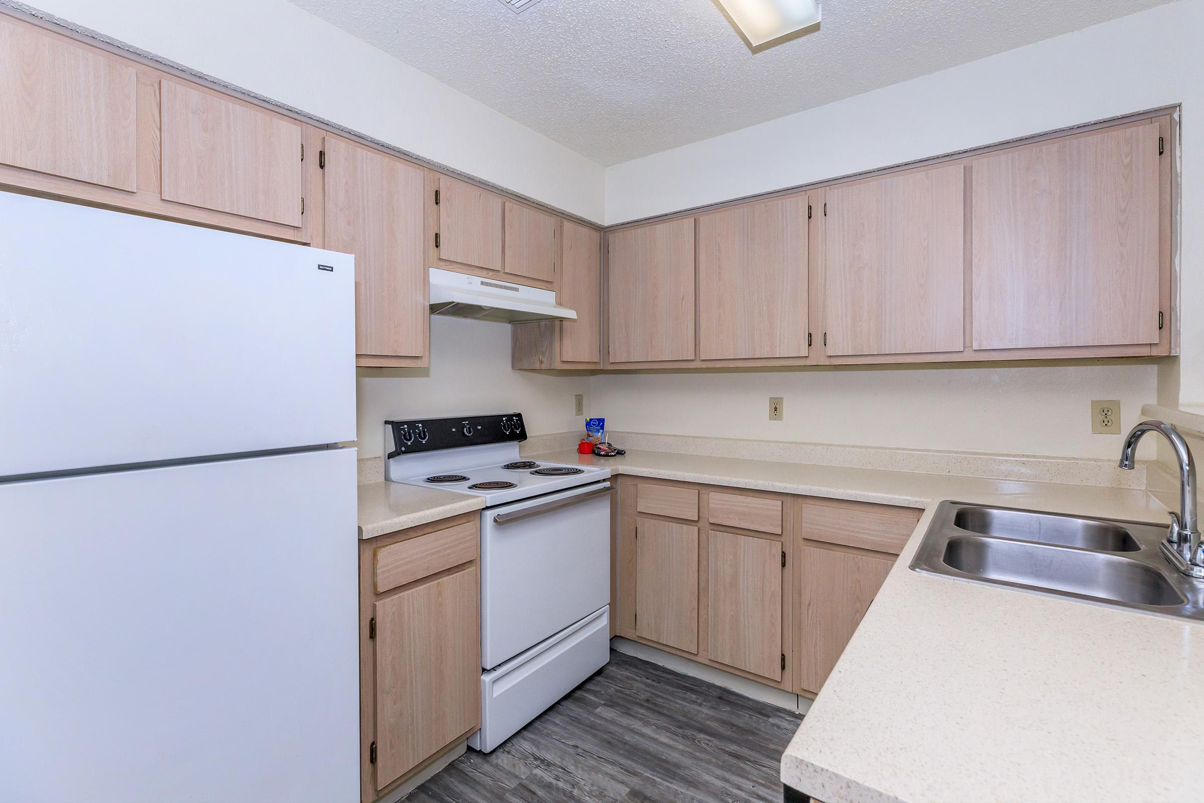 A clean, modern kitchen featuring light wood cabinetry, a white refrigerator, and a white stove with an oven. The countertops are a light color, and there’s a stainless steel sink. The kitchen is well-lit with a ceiling light, and the flooring appears to be wood-like laminate.