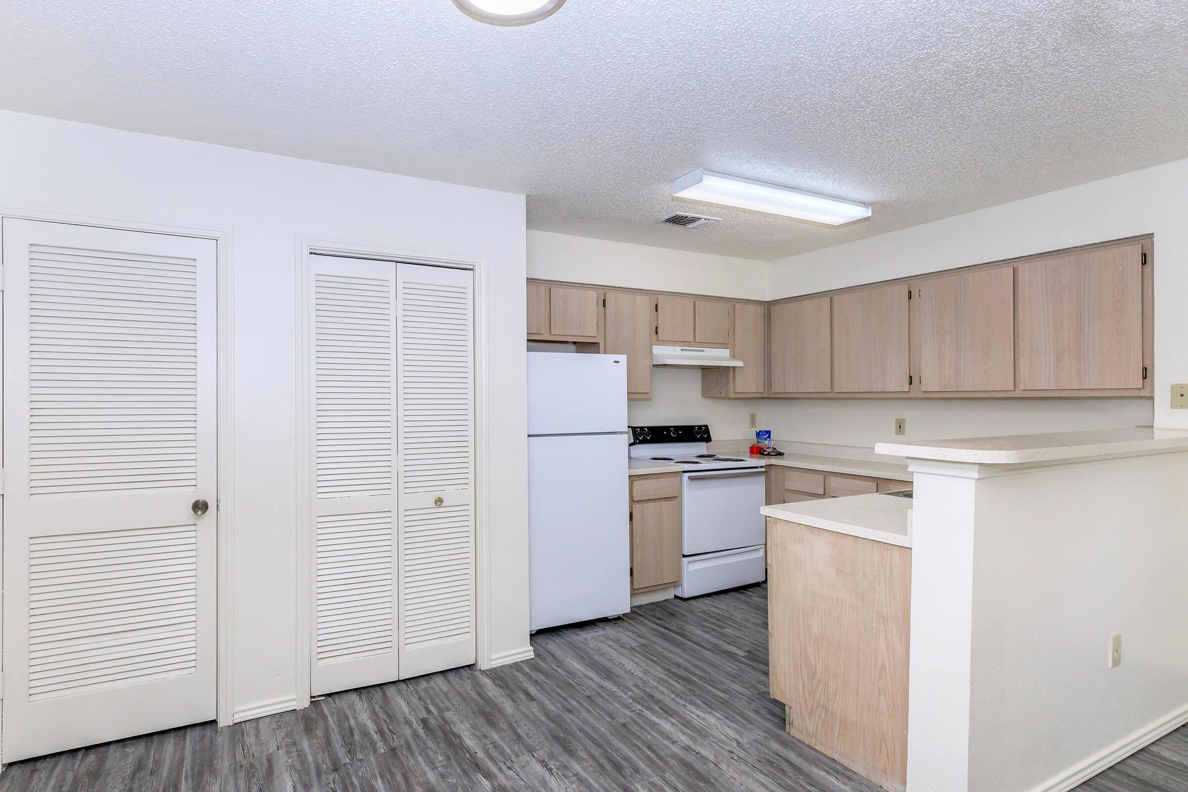 A modern kitchen featuring light wooden cabinets, white appliances including a refrigerator and stove, and a counter with a bar area. The room has a light-colored wall and a textured ceiling, with a door leading to a closet and modern flooring. Natural light illuminates the space.