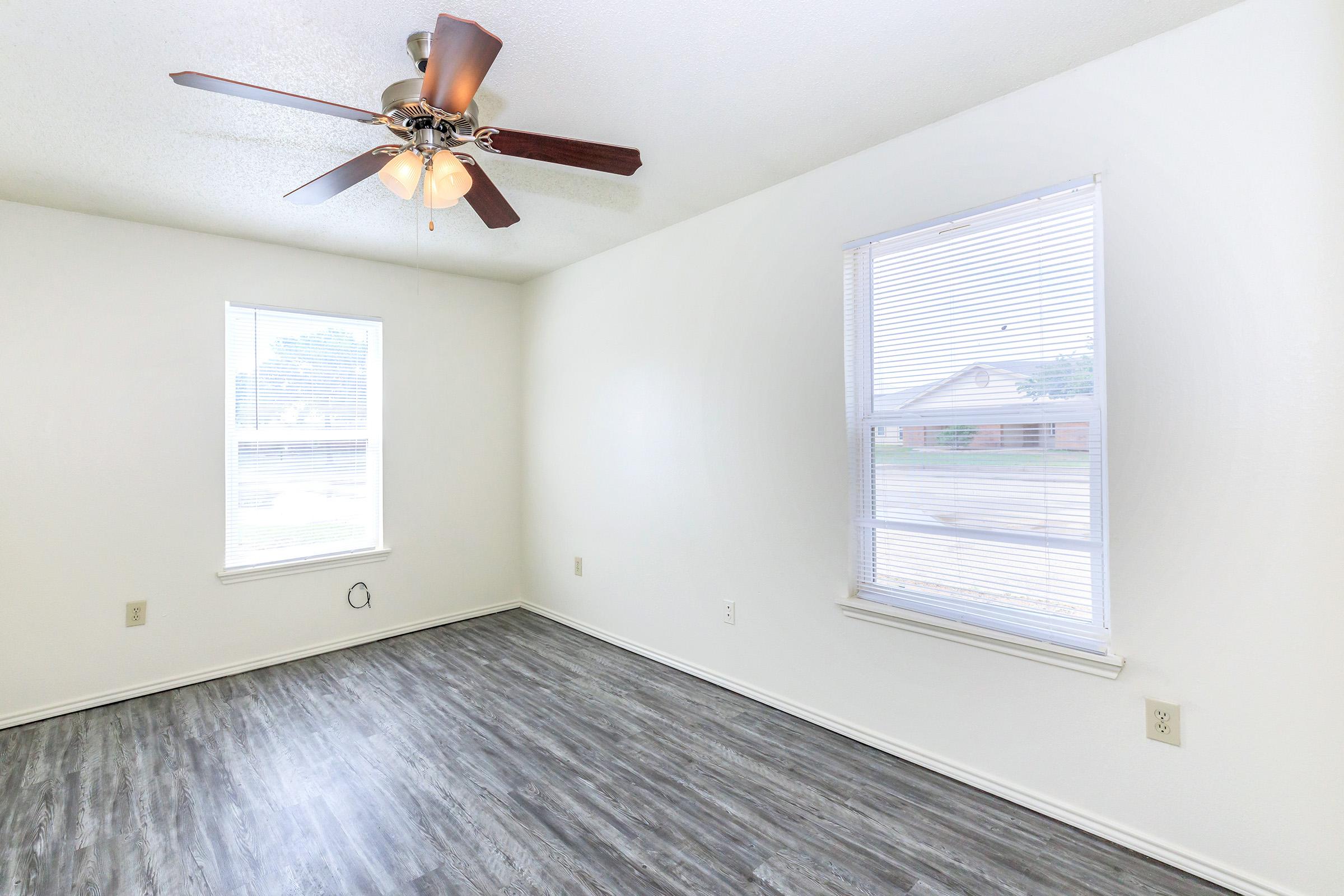 A spacious, empty room with light-colored walls and a modern ceiling fan with five bulbs. It features two windows with blinds, allowing natural light to enter, and a dark wood-like laminate floor. The room is clean and ready for furnishing.