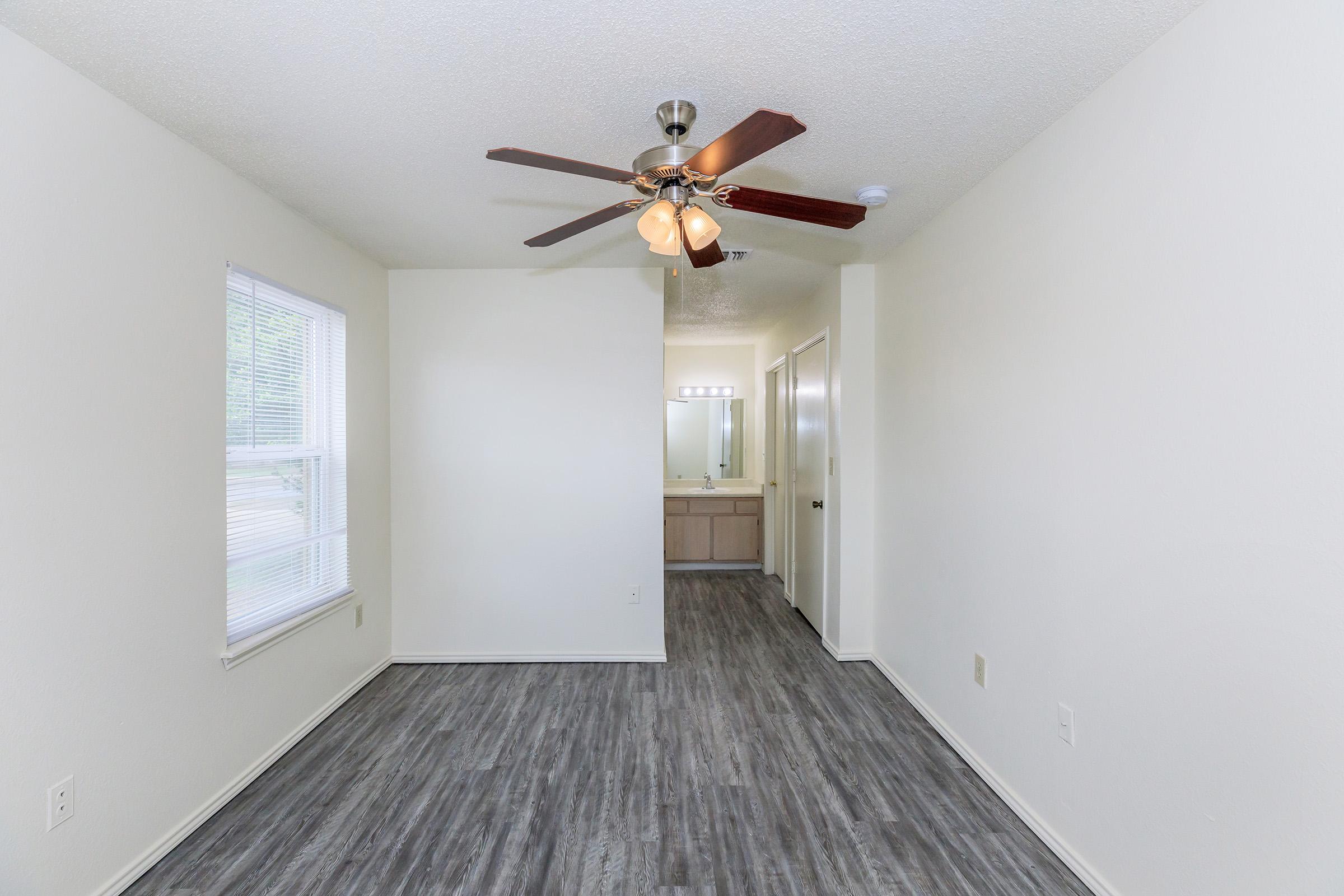 A light-colored room featuring a ceiling fan with wooden blades and a light fixture. The space has wood-like flooring and a large window allowing natural light. In the background, there is a doorway leading to a bathroom area, enhancing the open feel of the room. Walls are painted in neutral tones.