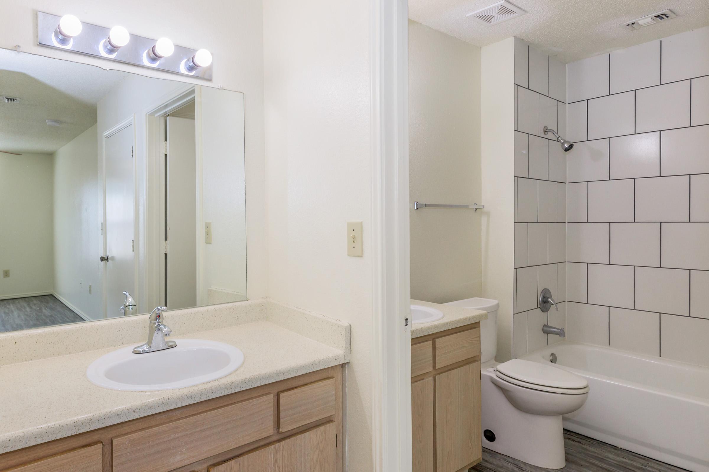 A well-lit bathroom featuring a double sink vanity with a mirror and modern lighting above. To the right, a shower and bathtub combo with white tiled walls. The floor has a light wood-like finish, and the walls are painted in neutral tones, creating a spacious and inviting atmosphere.