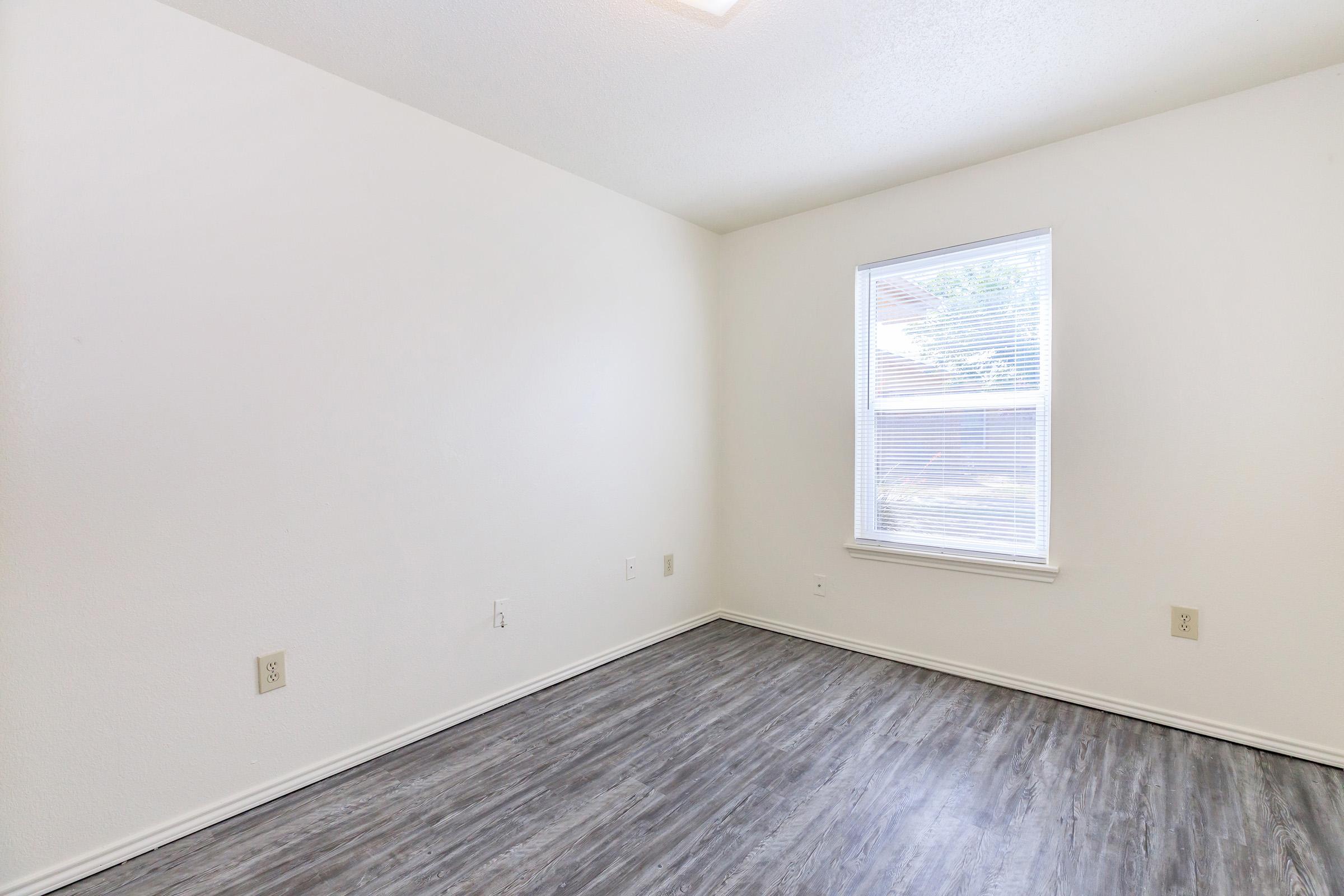 Empty room with light-colored walls and a large window letting in natural light. The floor is covered with a gray laminate, and there are no furnishings or decorations, creating a spacious and minimalistic appearance.