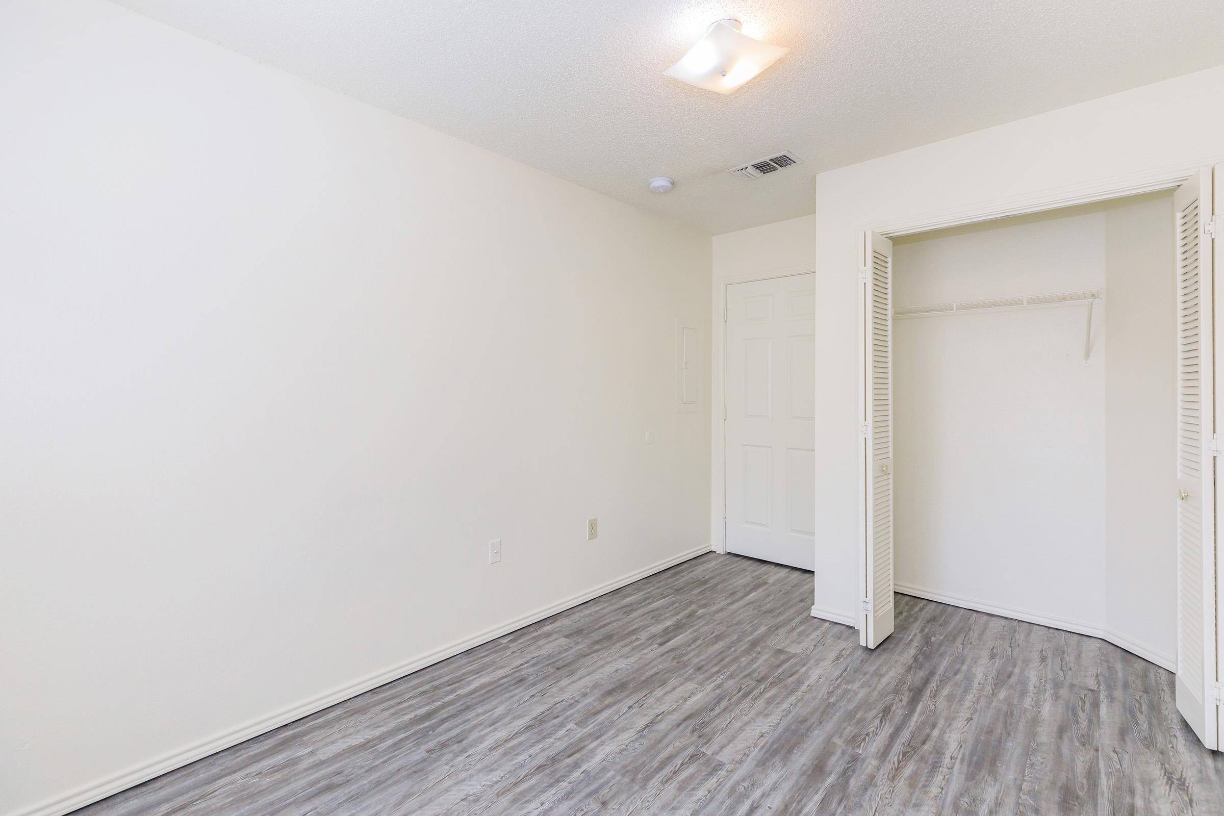 A spacious, empty room with light-colored walls and a wood-like floor. There is a bright ceiling light, a closed closet with sliding doors, and a door leading to another area. The overall ambiance is clean and inviting, suitable for various uses.