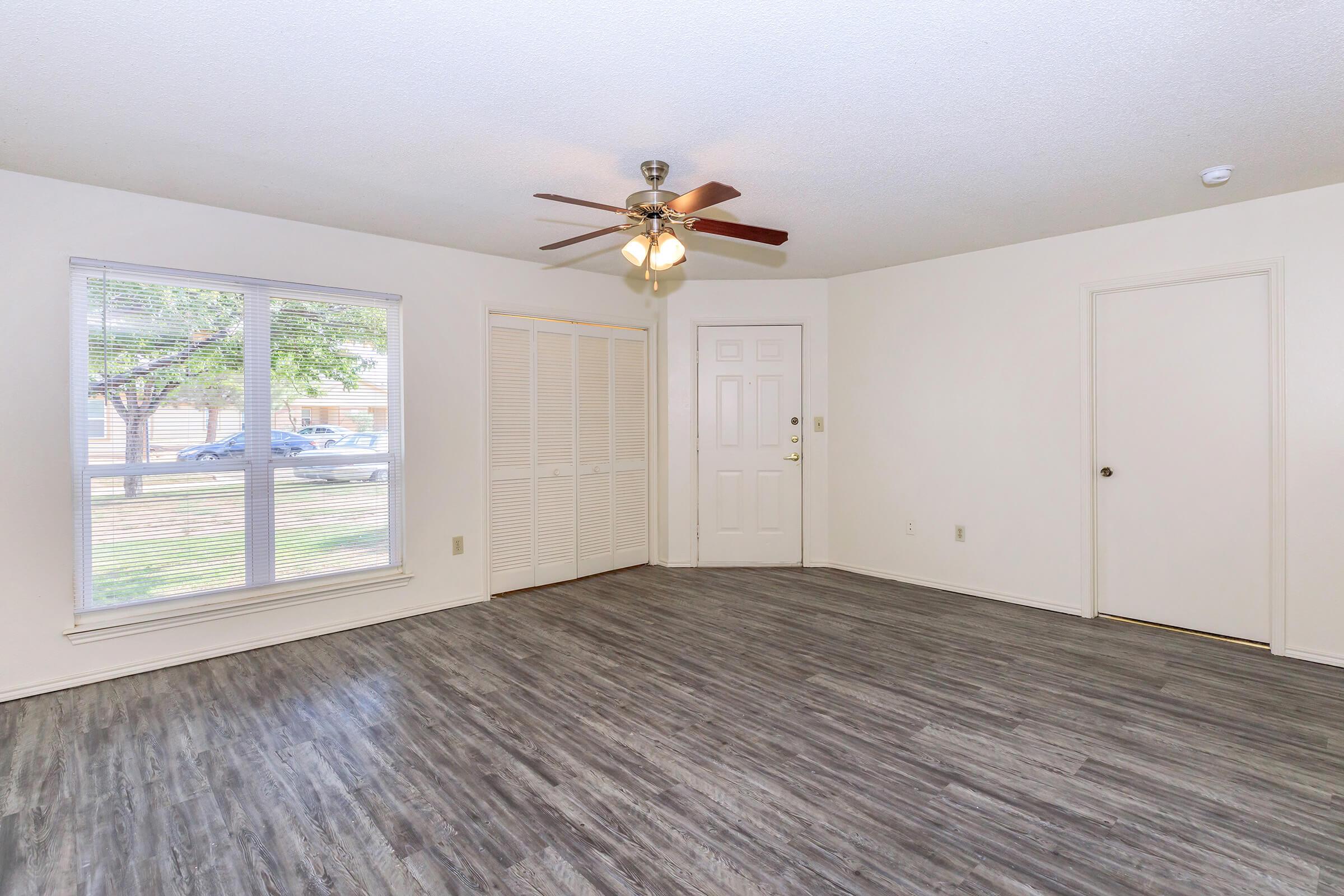 A spacious, empty living room with light-colored walls and a ceiling fan. There is a large window allowing natural light, a closet with sliding doors, and a front door. The flooring is a modern gray laminate, creating a clean, bright atmosphere.
