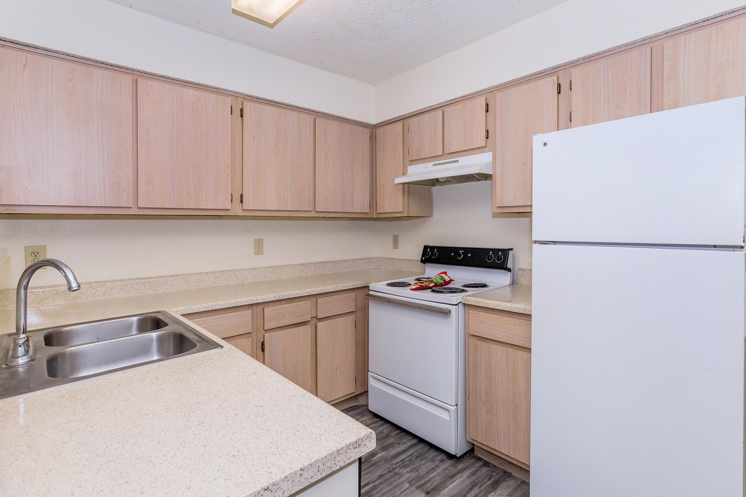 A modern kitchen featuring light-colored wooden cabinets, a white refrigerator, a white stove with an overhead range hood, and a double sink with a modern faucet. The countertops are light-colored and smooth, and the flooring appears to be a wood-like laminate. Fresh ingredients are placed on the counter.