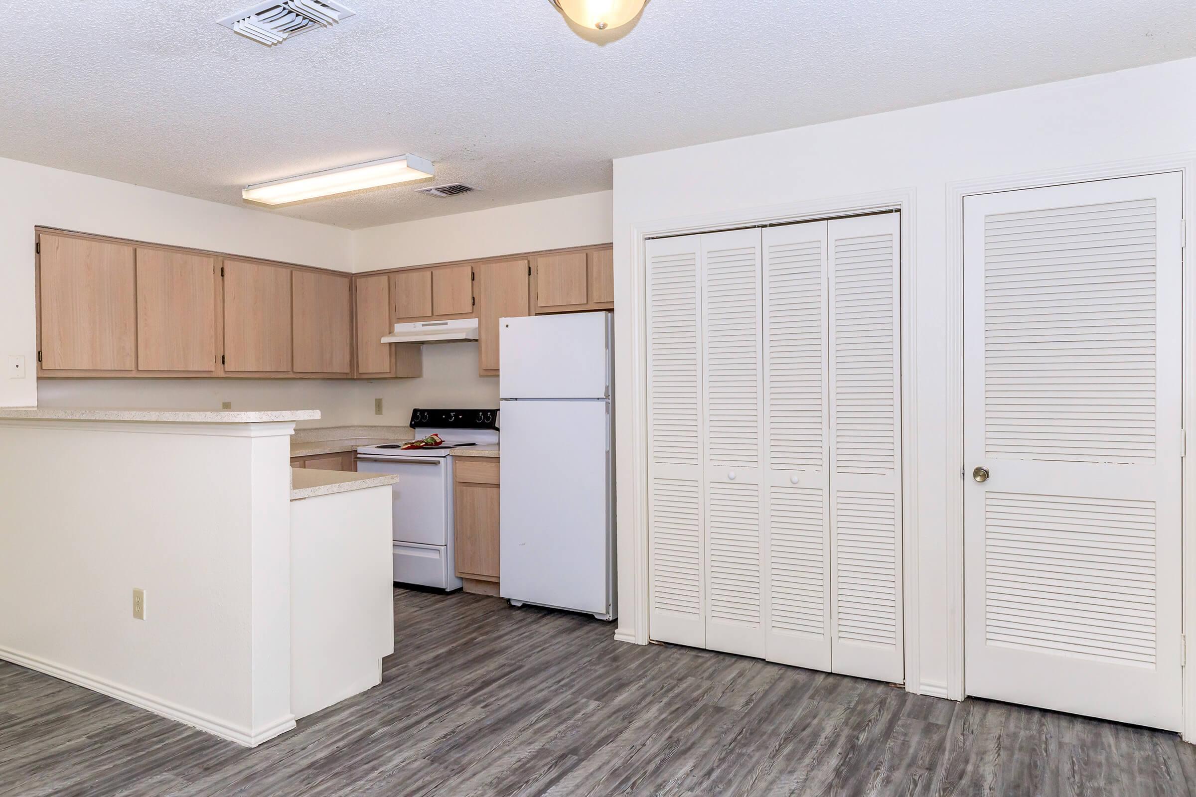 A bright, modern kitchen featuring light wood cabinets, white appliances, and a spacious layout. The room includes a refrigerator, stove, and an island with a countertop. There's a closet with bi-fold doors on the right, and the flooring is a sleek, dark laminate. Natural light enters from overhead fixtures.