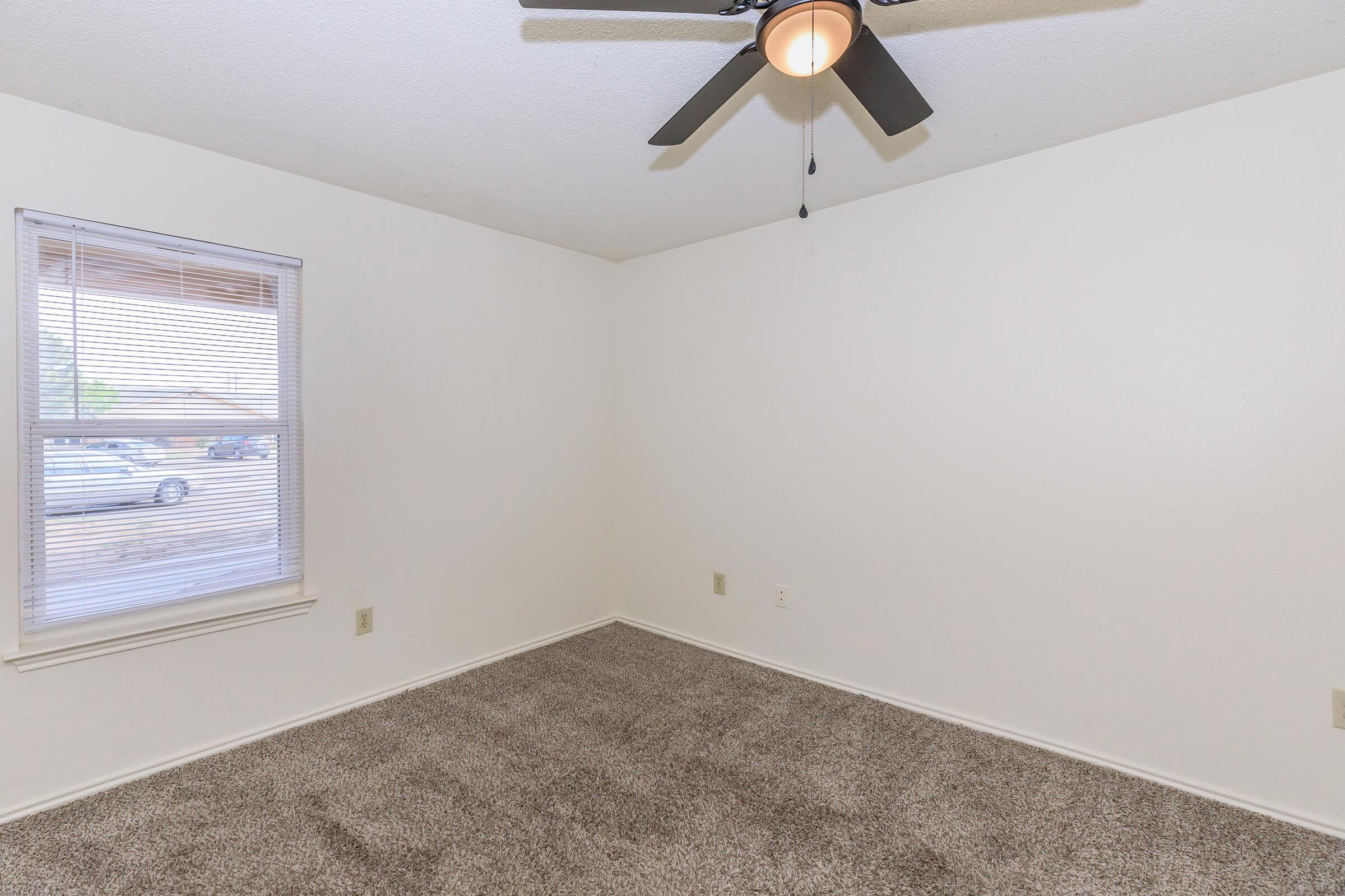 A vacant room featuring beige carpet, a ceiling fan, and a window with blinds. The walls are painted white, creating a bright and airy atmosphere. The room is minimalistic, with no furniture present, highlighting its spaciousness and potential for personalization. Natural light enters through the window.